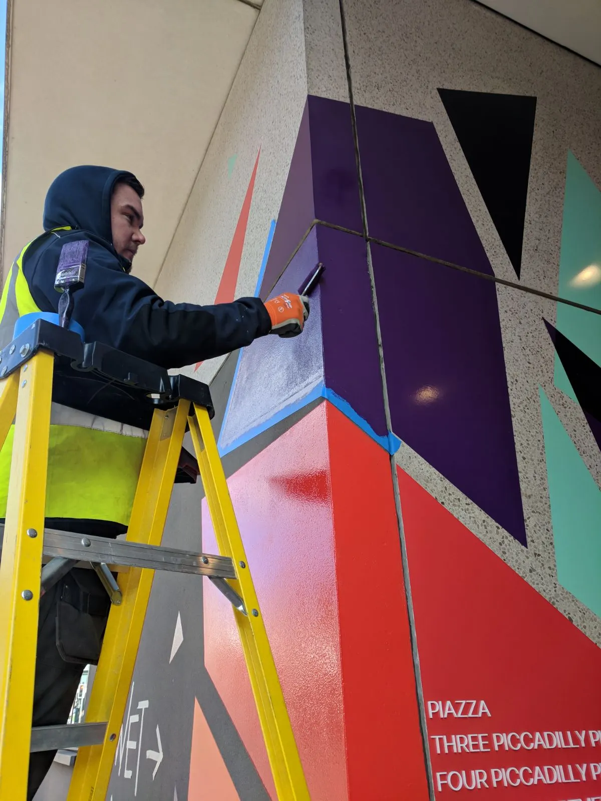 Person on a ladder painting a colorful geometric mural on a building wall.
