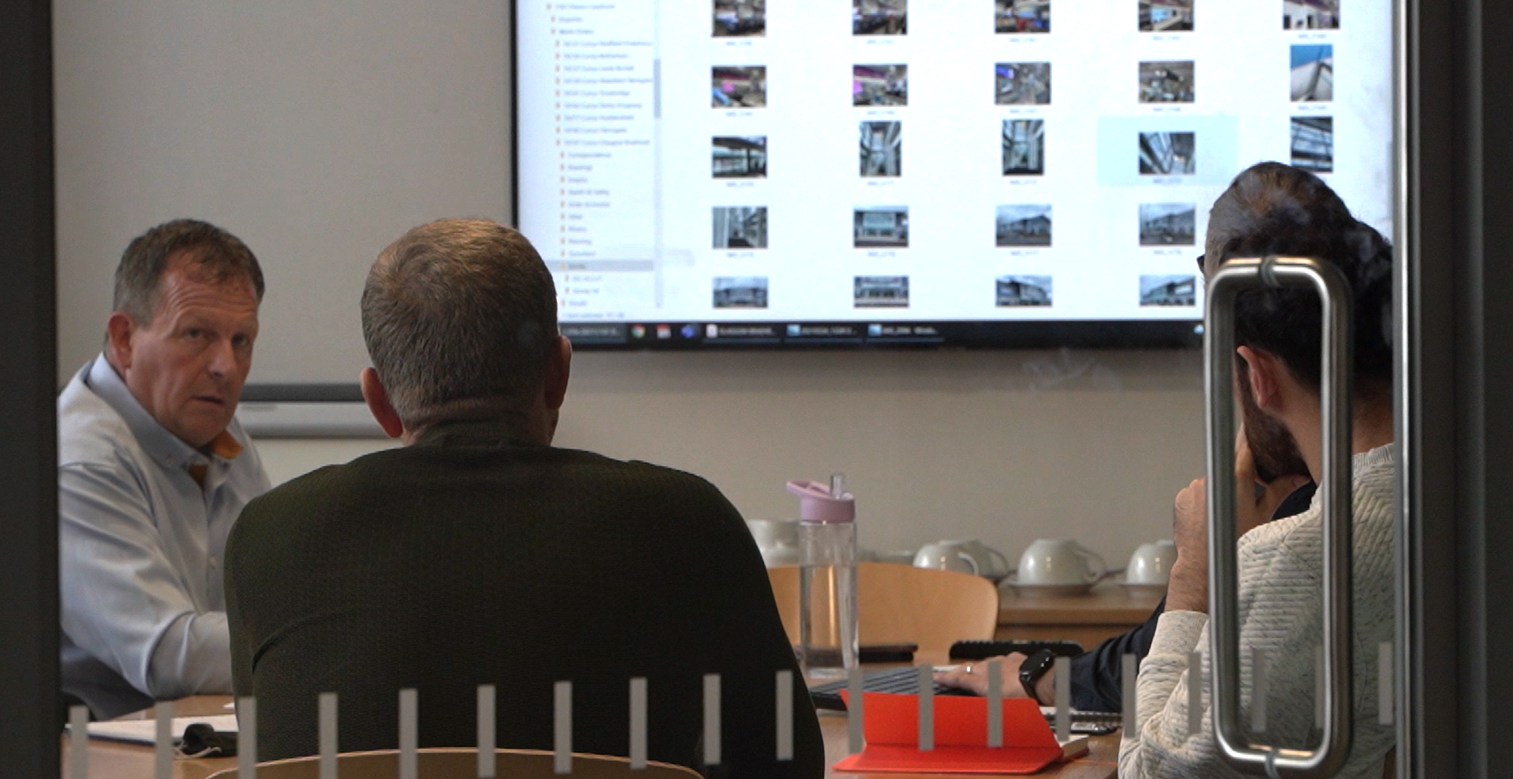 Business meeting in a modern office room with four people seated at a table. A large screen displays a grid of thumbnail images, possibly for a presentation. Notebooks and hard hats are visible on the table, indicating a construction or engineering context. Glass walls with vertical stripes add a contemporary touch.