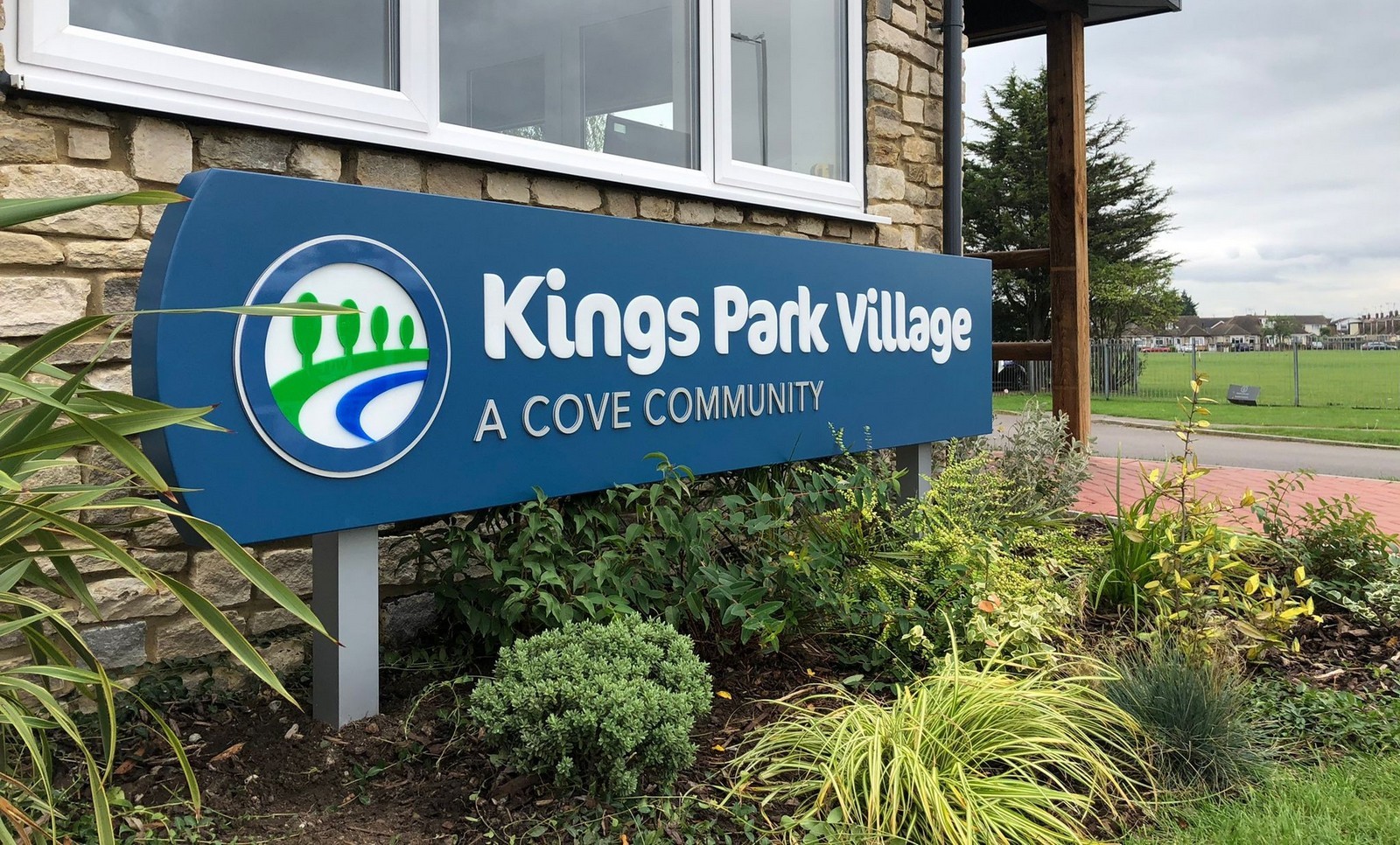 Sign reading "Kings Park Village: A Cove Community" surrounded by plants and grass.