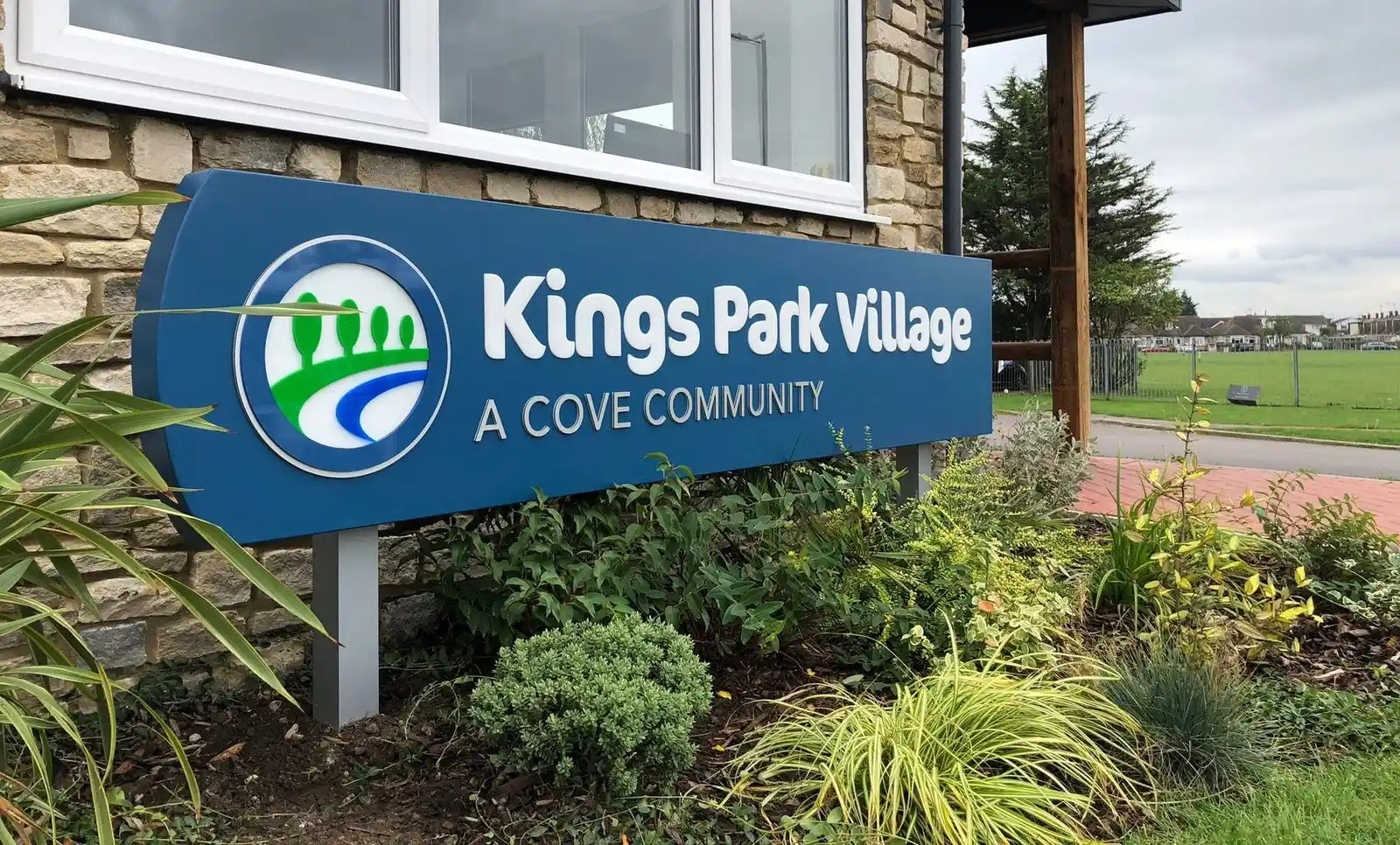 Sign reading "Kings Park Village: A Cove Community" surrounded by plants and grass.