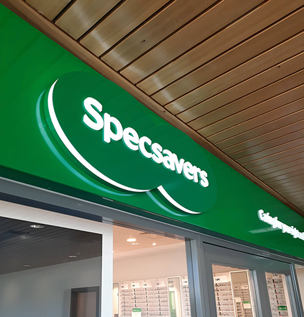Green Specsavers sign with white lettering on a storefront under a wooden ceiling.