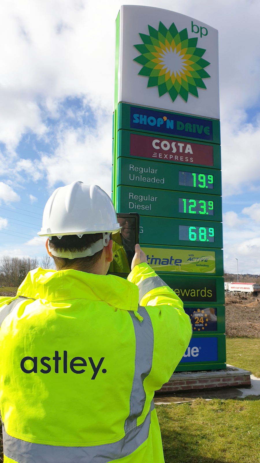 A worker in a high-visibility jacket labeled "astley" inspects a BP fuel station sign displaying fuel prices: Regular Unleaded at 119.9, Regular Diesel at 123.9, and Ultimate Diesel at 168.9. The sign also features branding for Costa Express.