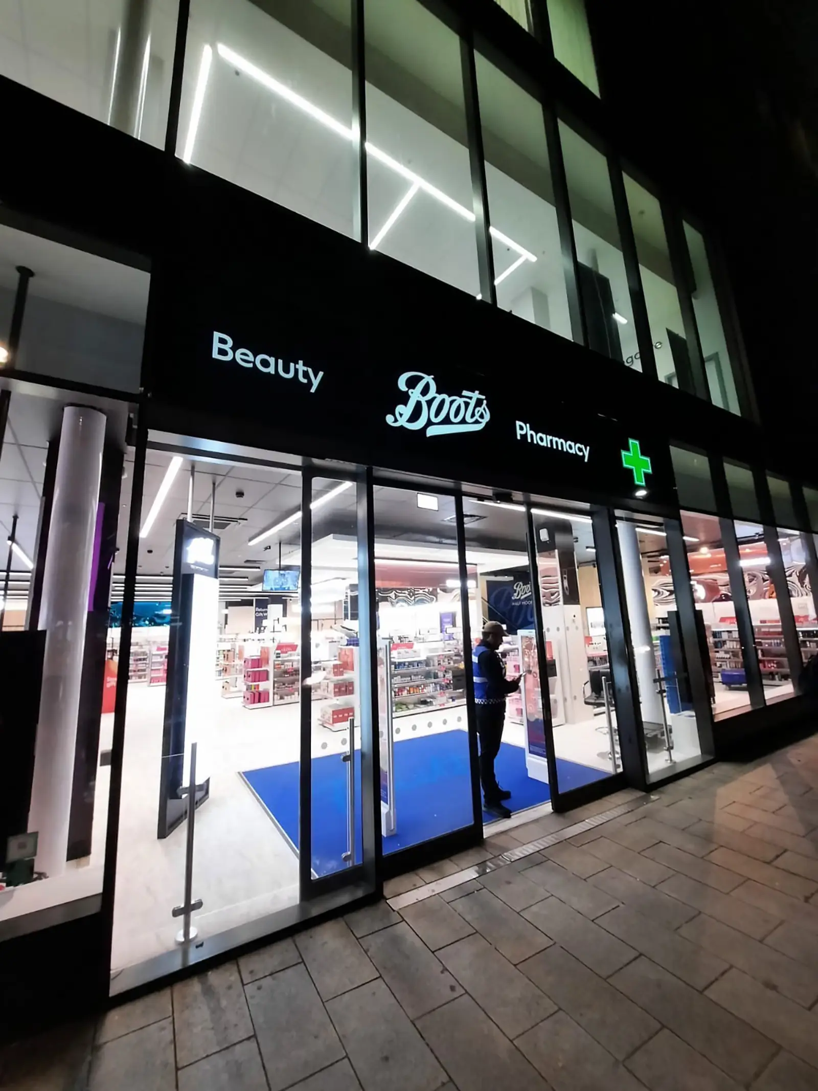 Exterior of a Boots store at night with illuminated signs and a person inside.