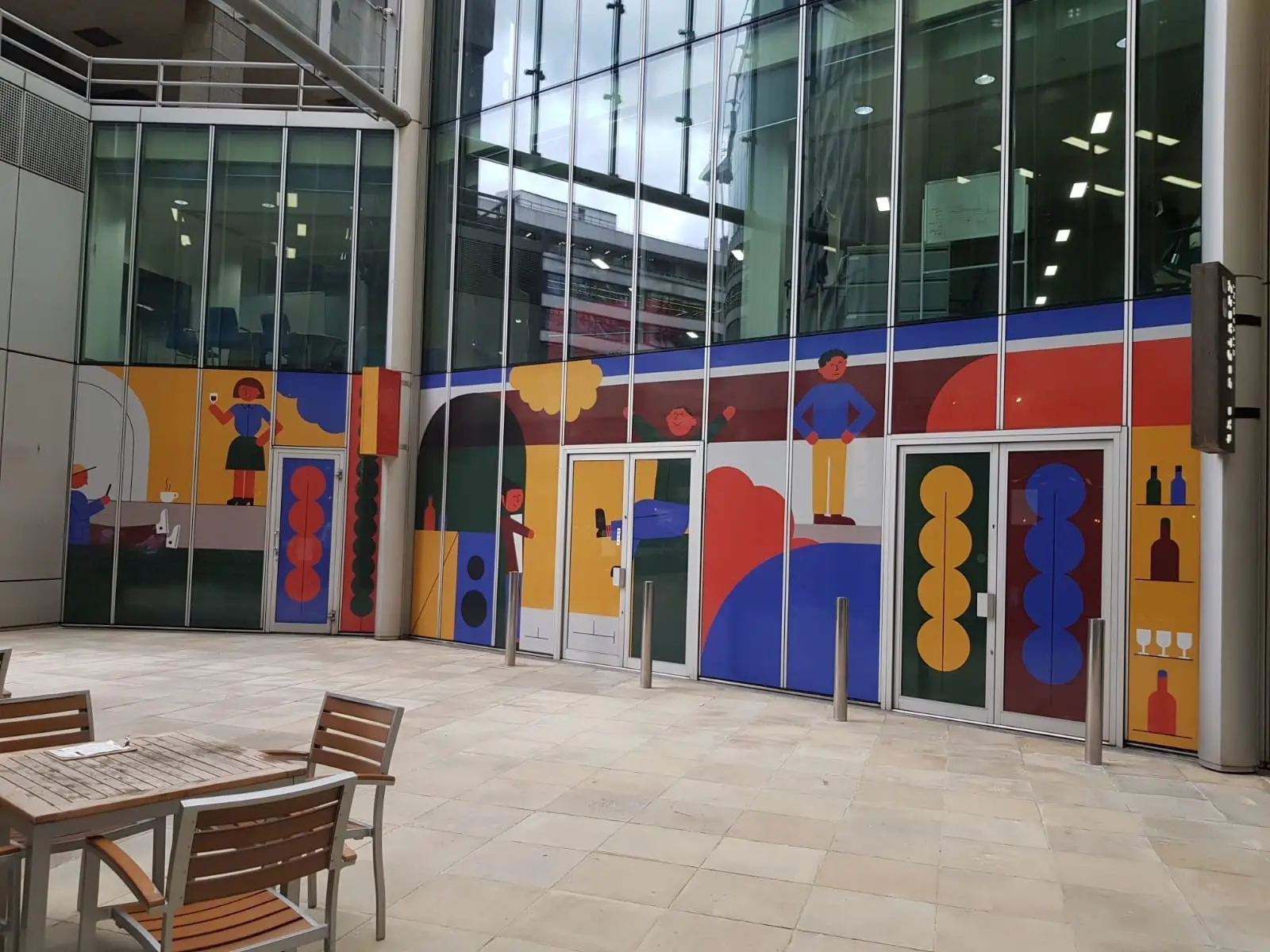 Colorful mural with abstract figures and shapes on a glass building facade near wooden tables.