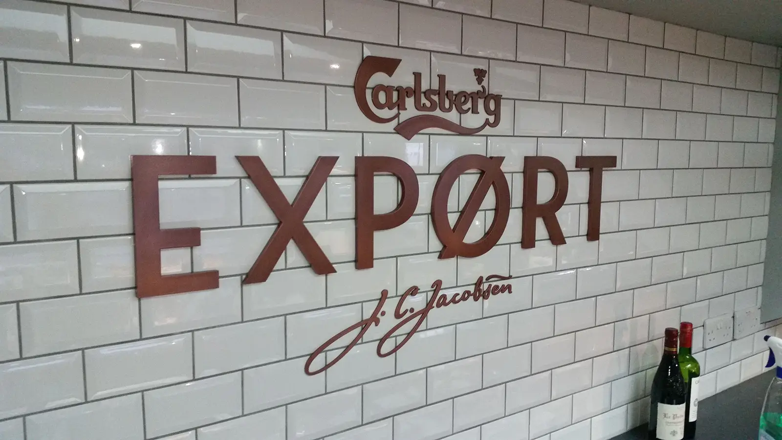 Carlsberg Export logo on a white tiled wall with bottles on a counter below.