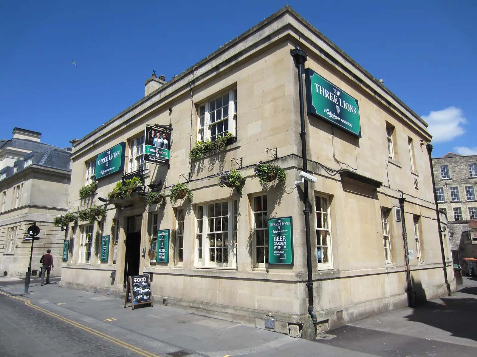 A corner pub with hanging plants and signs reading "The Three Lions" on a sunny day.