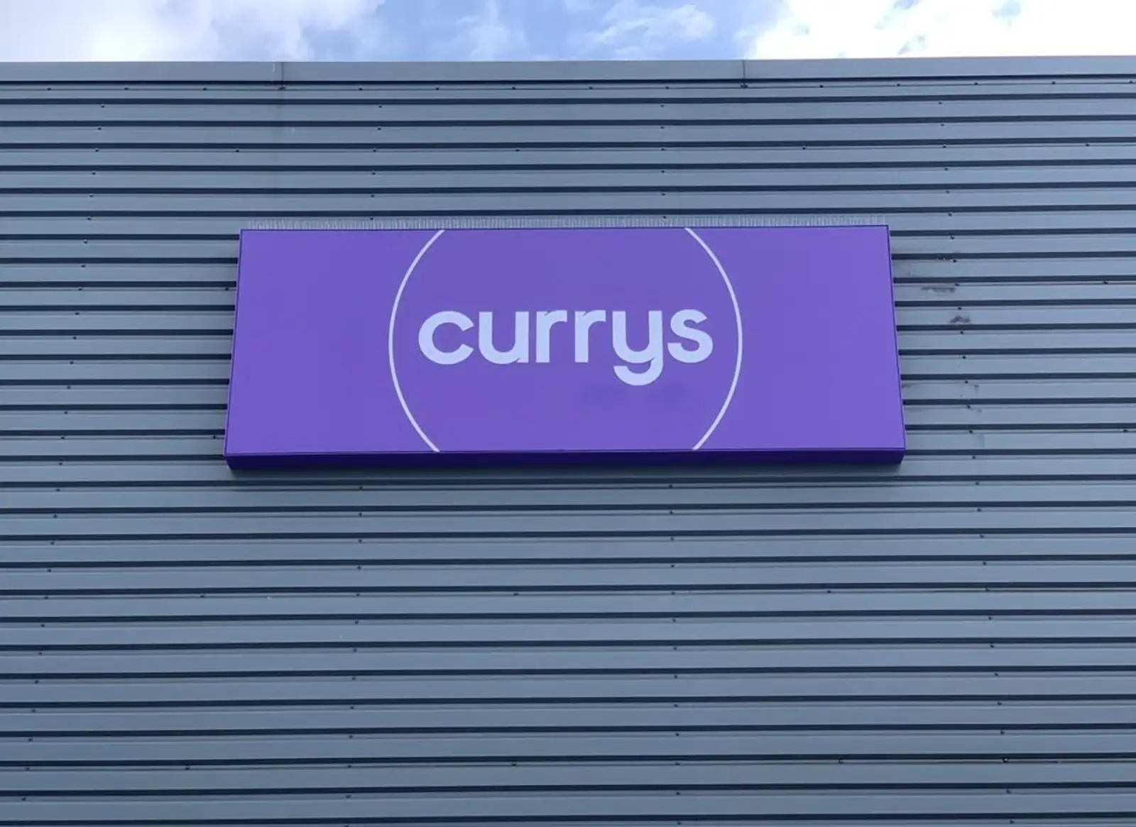 A purple sign with "currys" in white letters on a corrugated metal building.