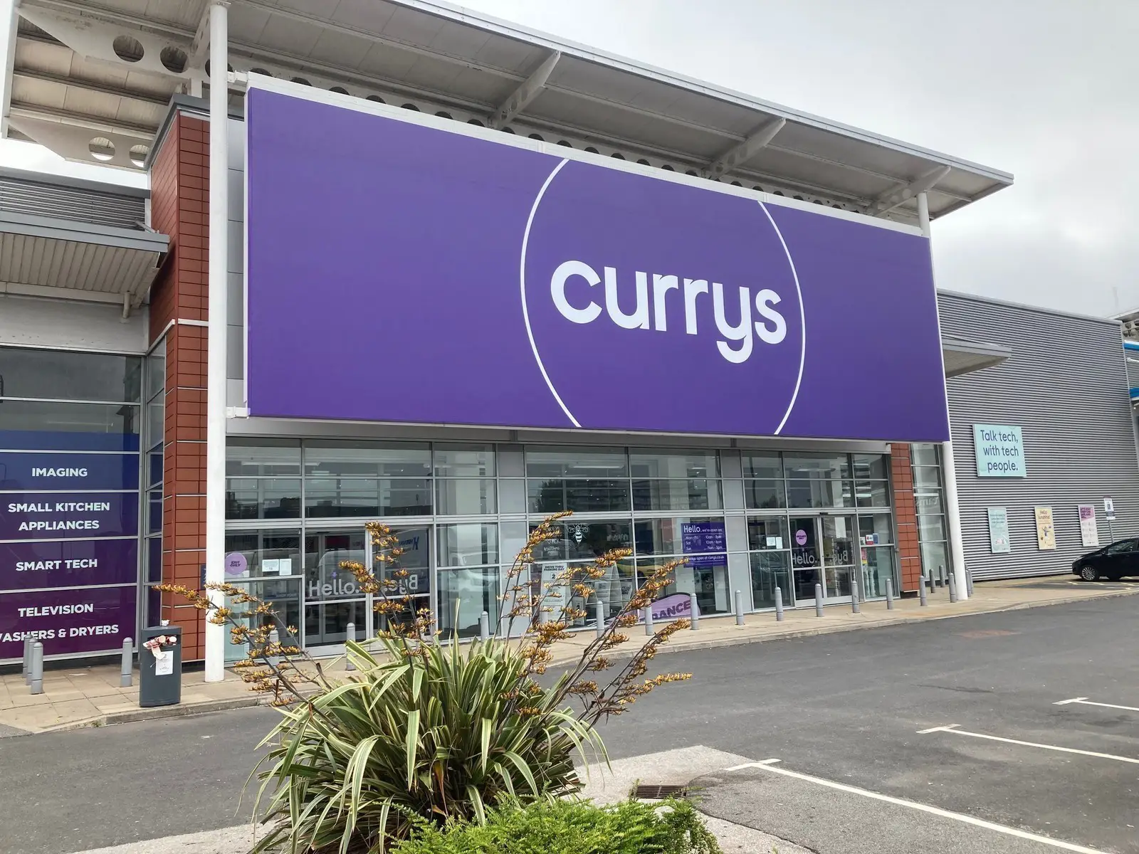 Large Currys storefront with a purple sign and a plant in the foreground.