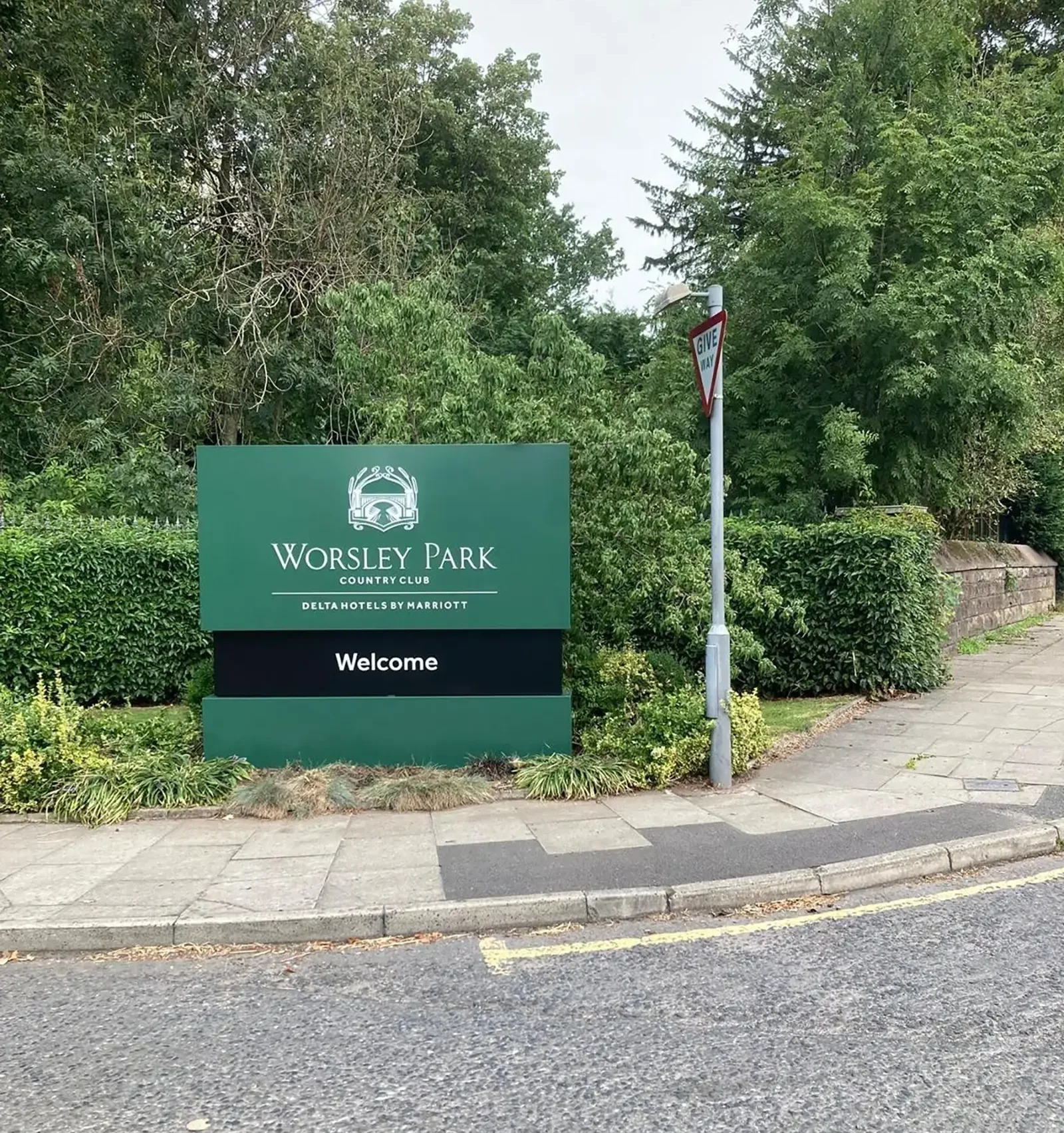 Green sign for Worsley Park Country Club surrounded by trees and bushes near a sidewalk.