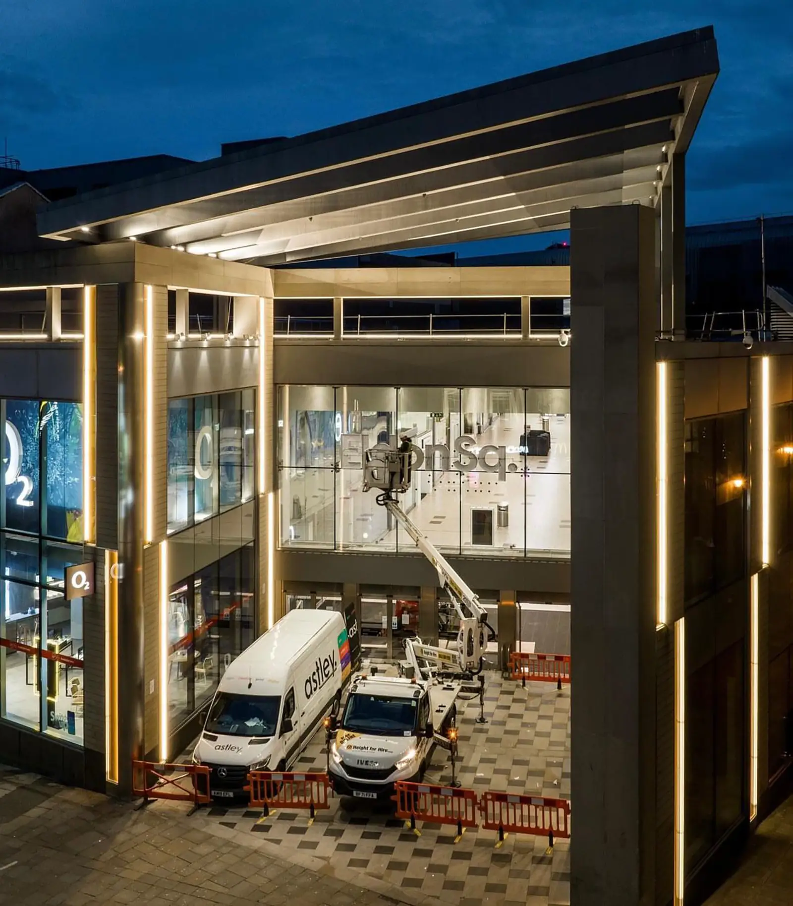 Modern building with illuminated facade, two parked service vehicles, and a cherry picker at dusk.