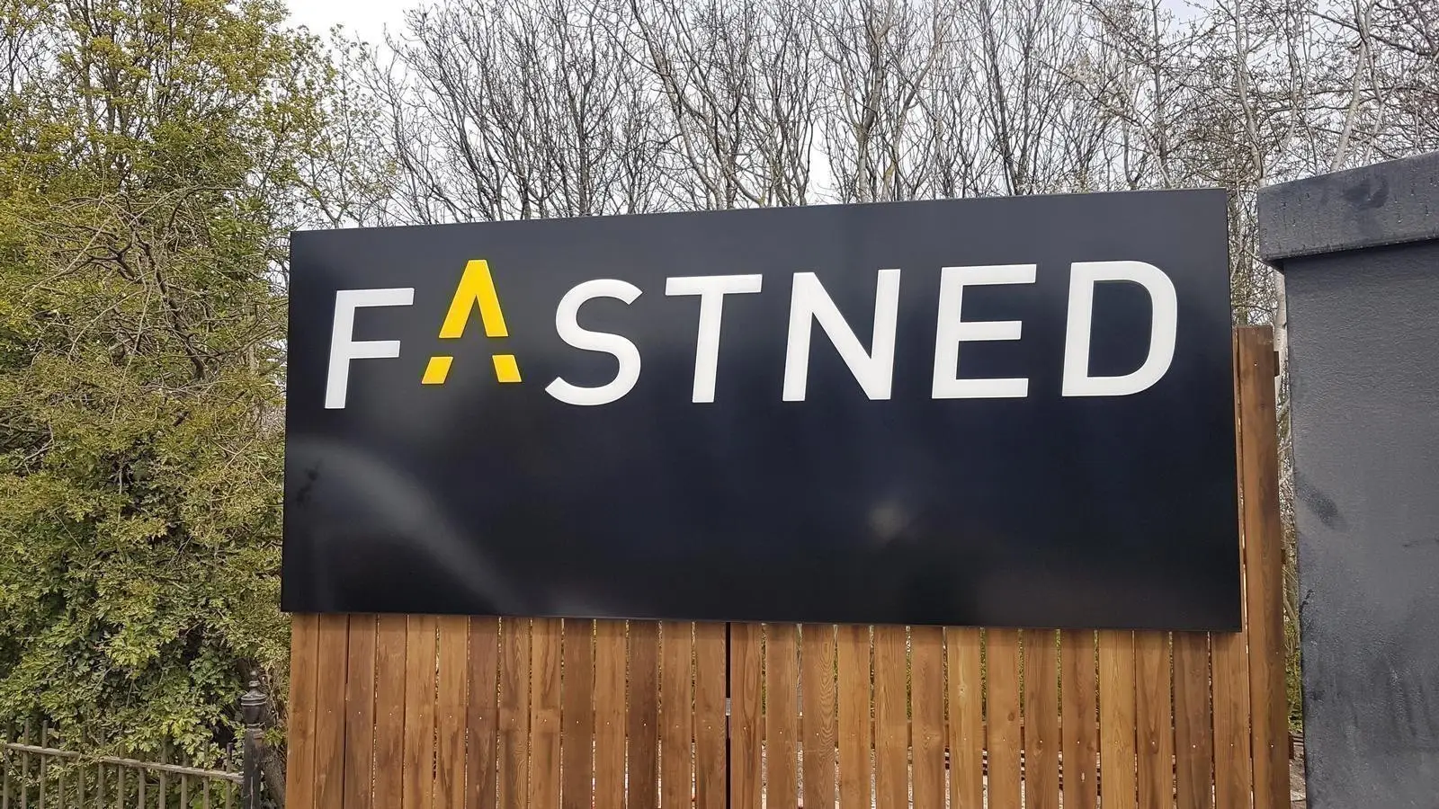 A black sign with "FASTNED" in white and yellow letters, surrounded by trees and fencing.