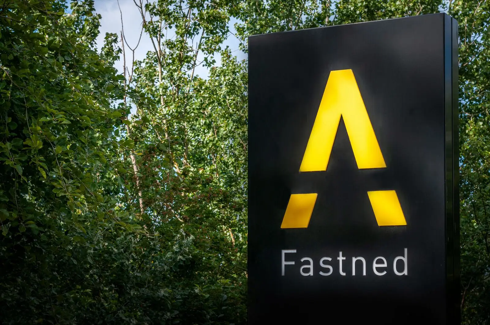 Black sign with a yellow stylized "A" and "Fastned" text, surrounded by green trees.