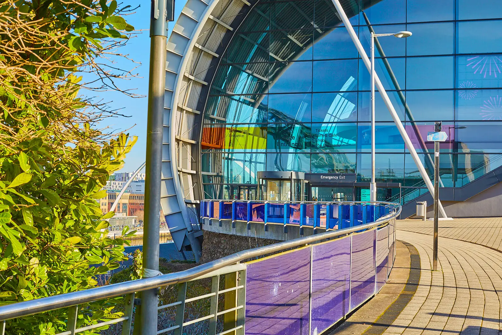 Curved modern glass building with colorful panels and a purple railing, surrounded by greenery.