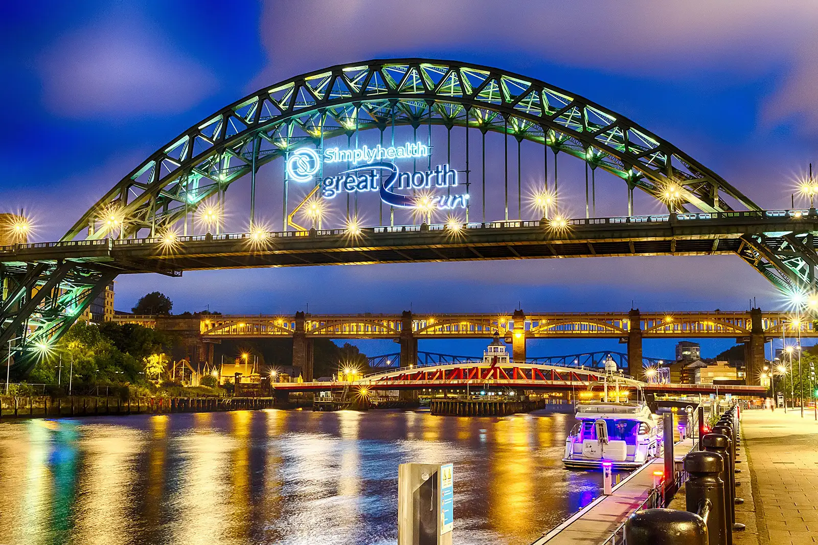 Illuminated bridge over a river at night with reflections and boats docked along the shore.