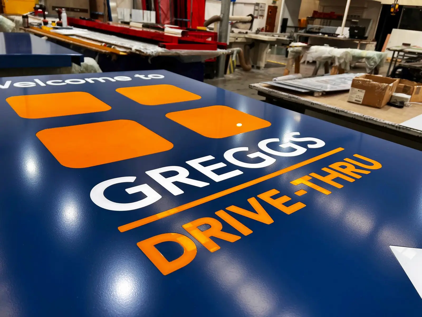 A blue sign with "Welcome to GREGGS DRIVE-THRU" in white and orange letters.
