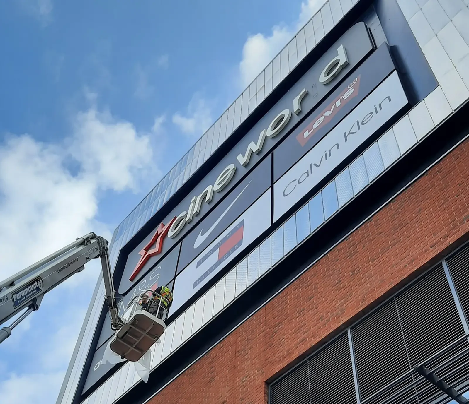 Workers on a lift install a large "Cineworld" sign on a building exterior.