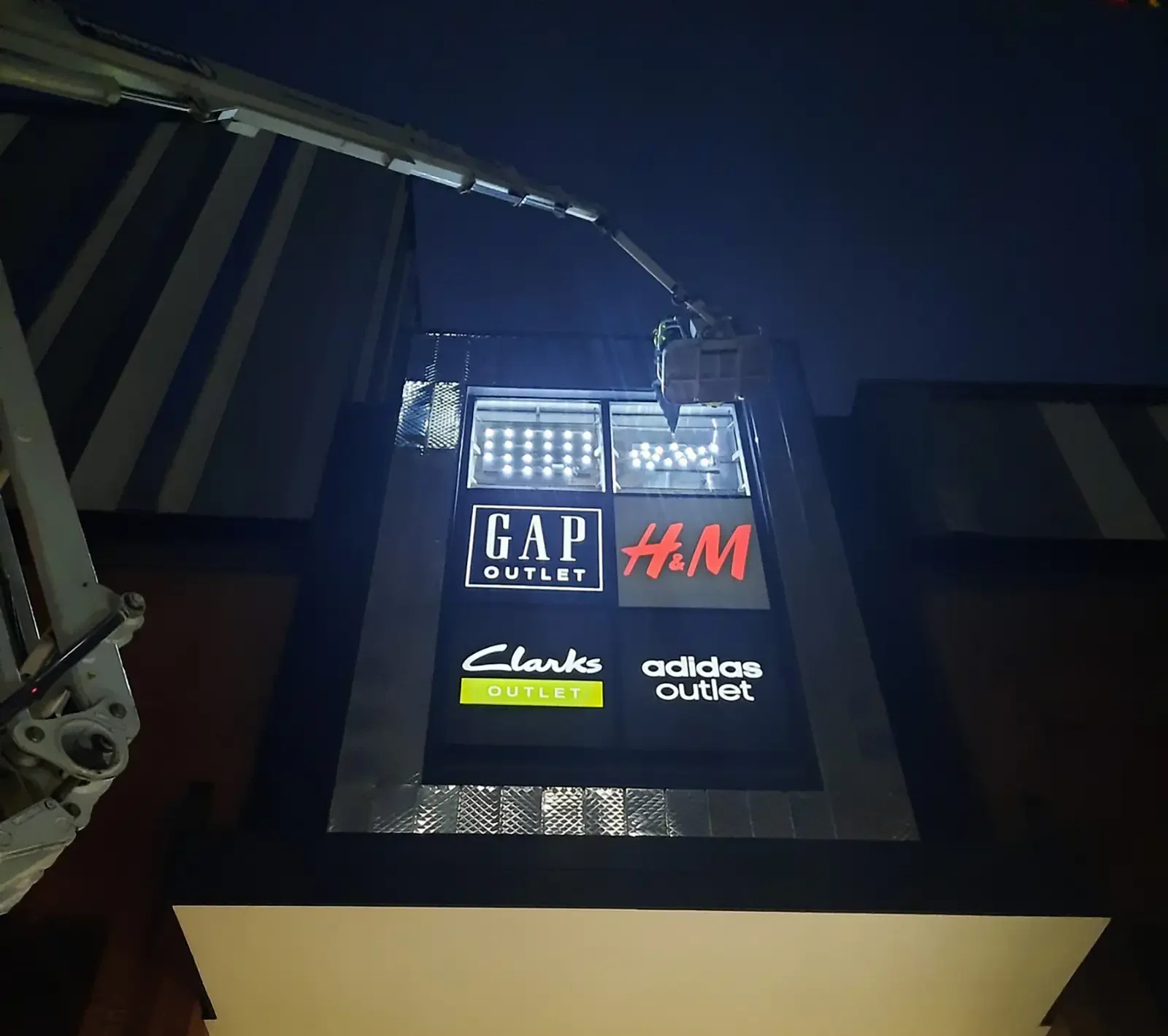 A worker in a lift repairs a sign displaying Gap, H&M, Clarks, and Adidas logos.