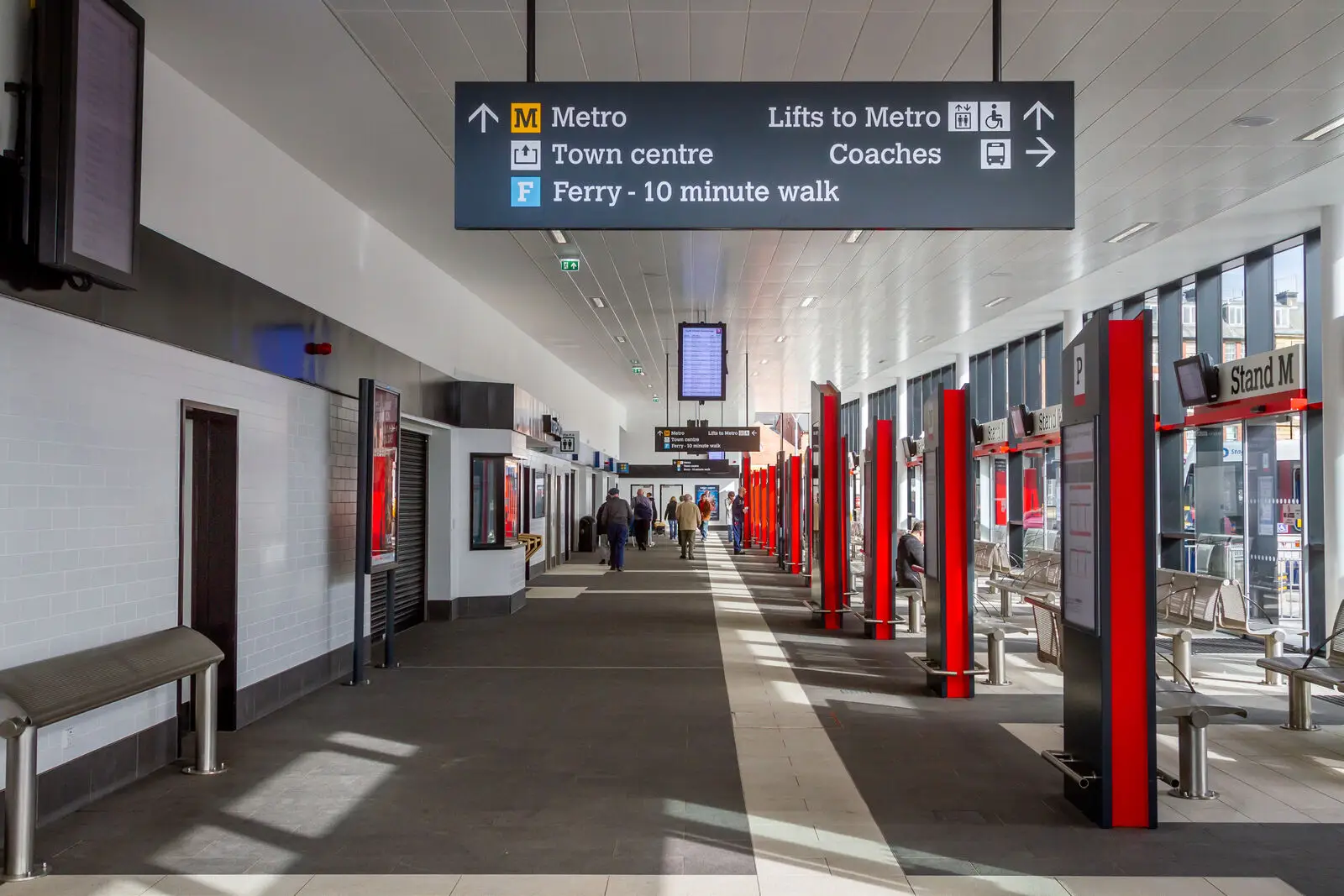 A bus station with red pillars, seating areas, and directional signs hanging from the ceiling.