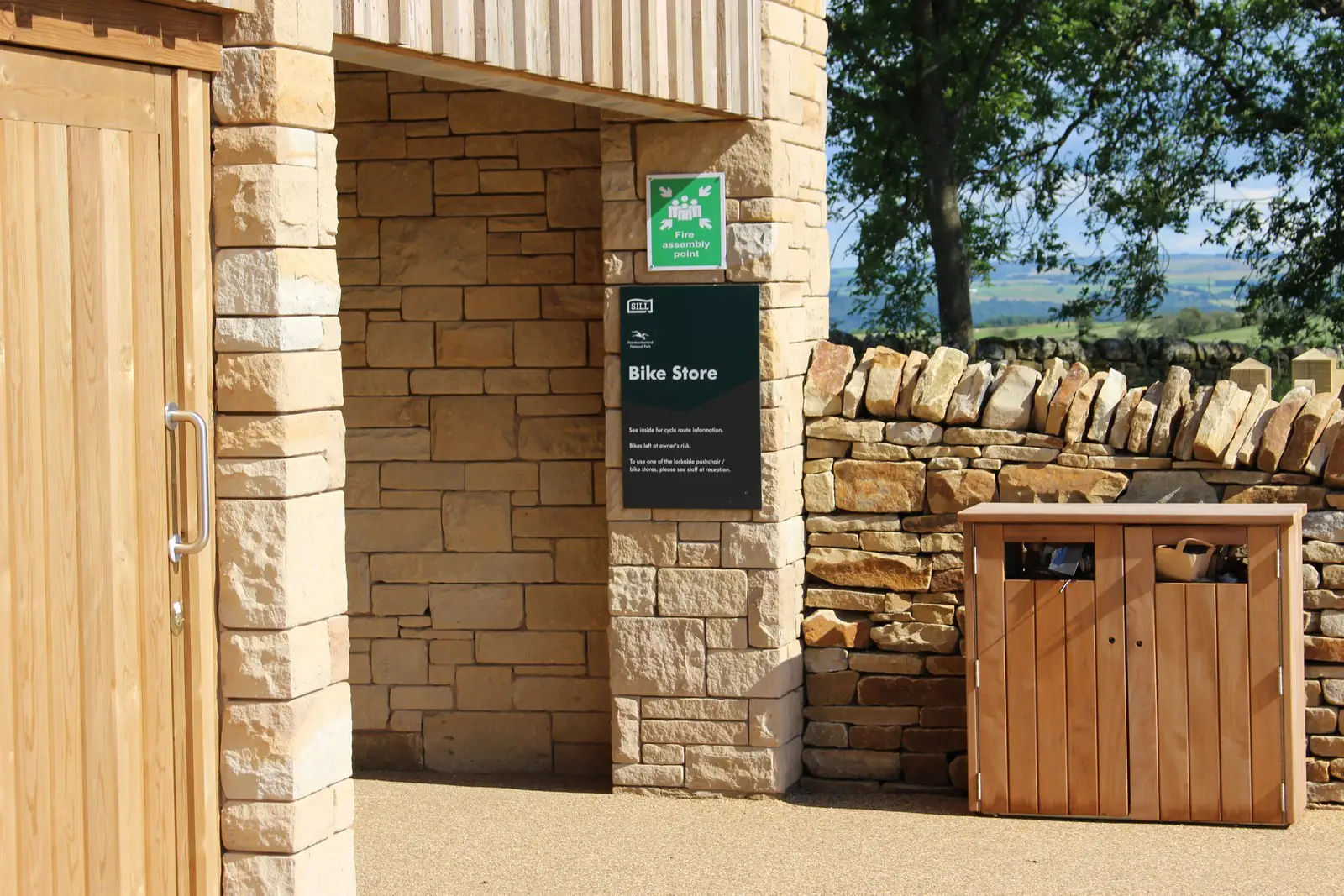 Stone building entrance with a "Bike Store" sign, wooden door, and wooden bin nearby.