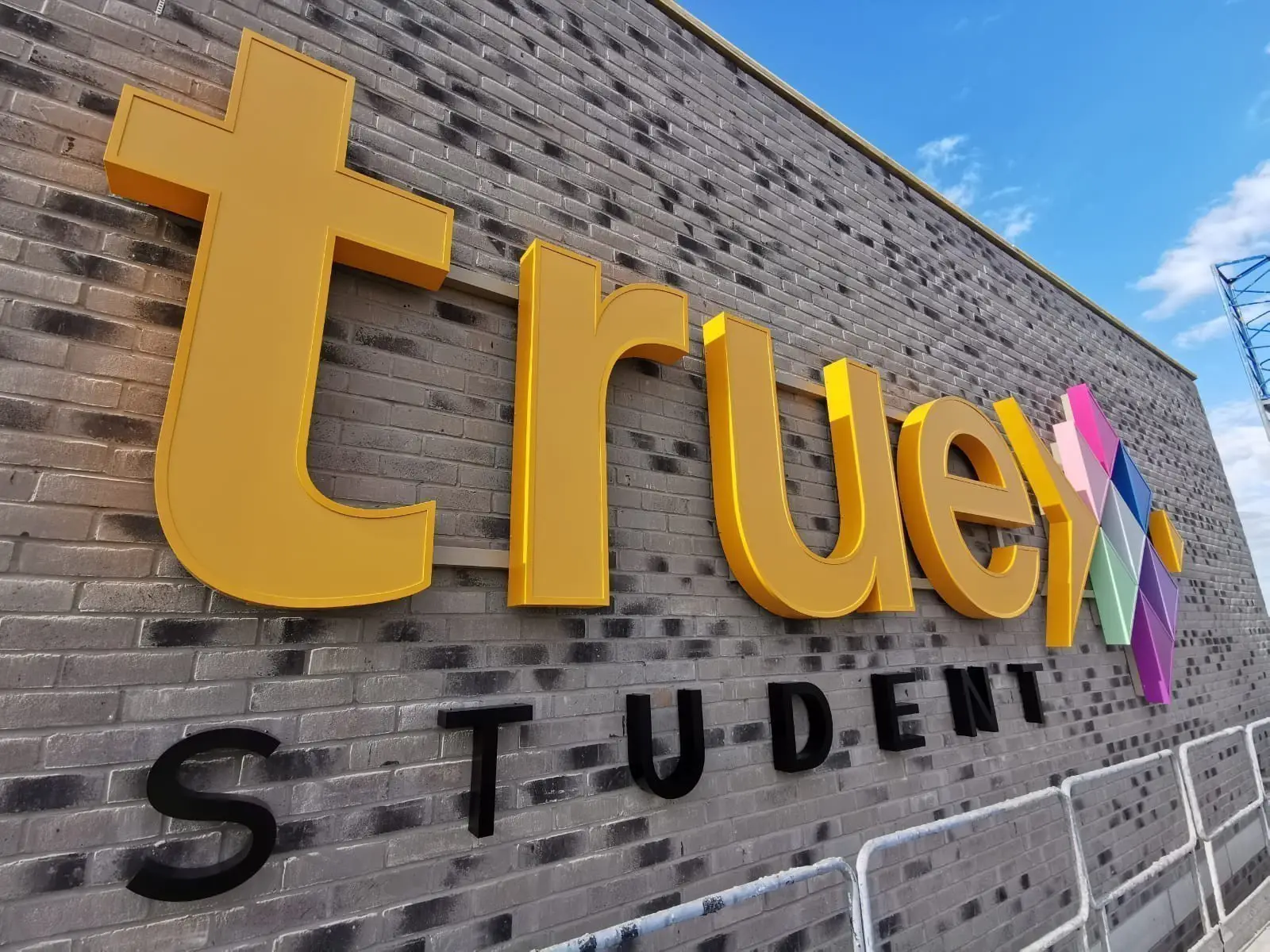 Large yellow "true" sign with colorful geometric shapes on a brick wall under a blue sky.