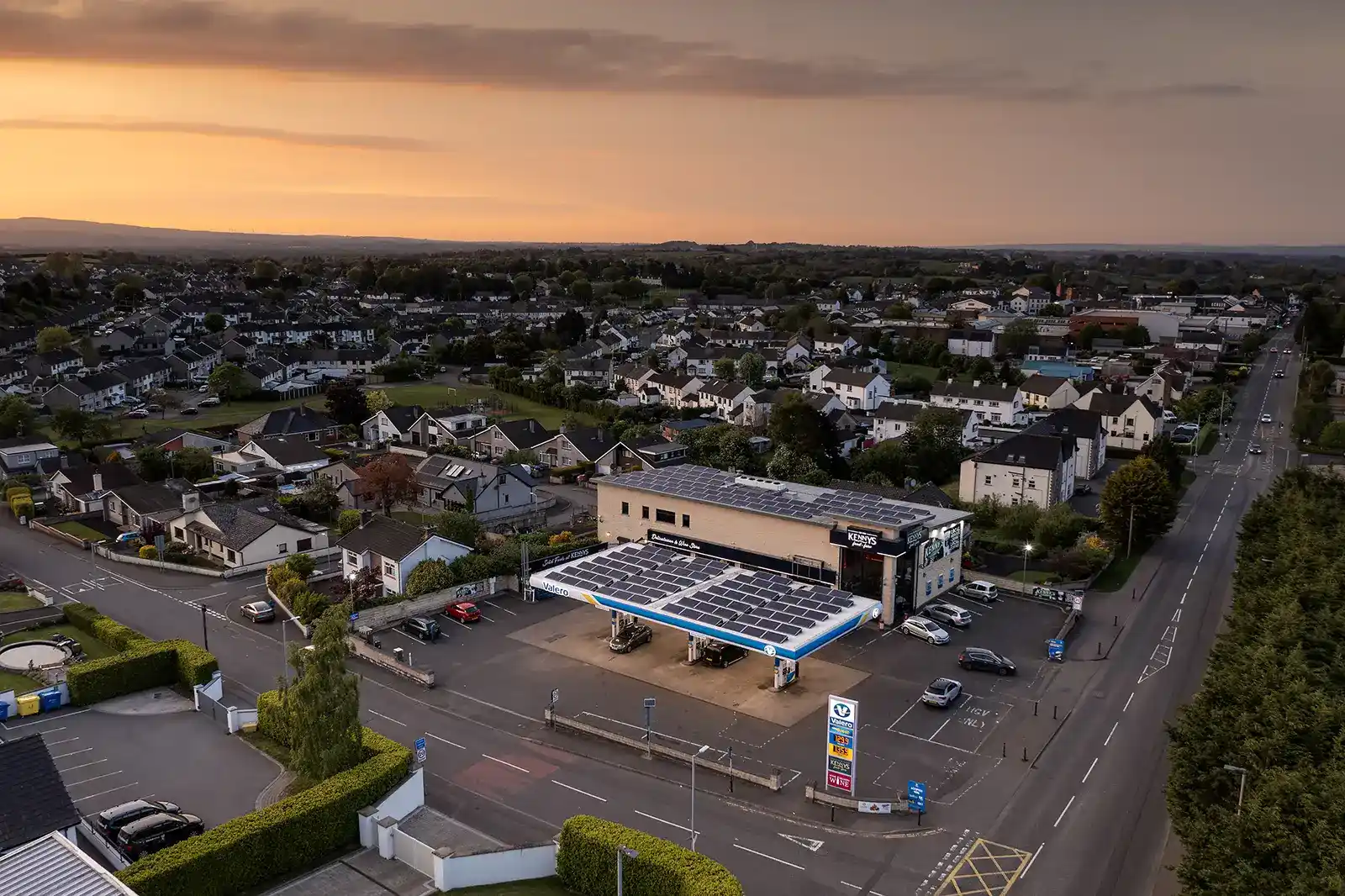 Aerial view of a gas station with solar panels, surrounded by houses at sunset.