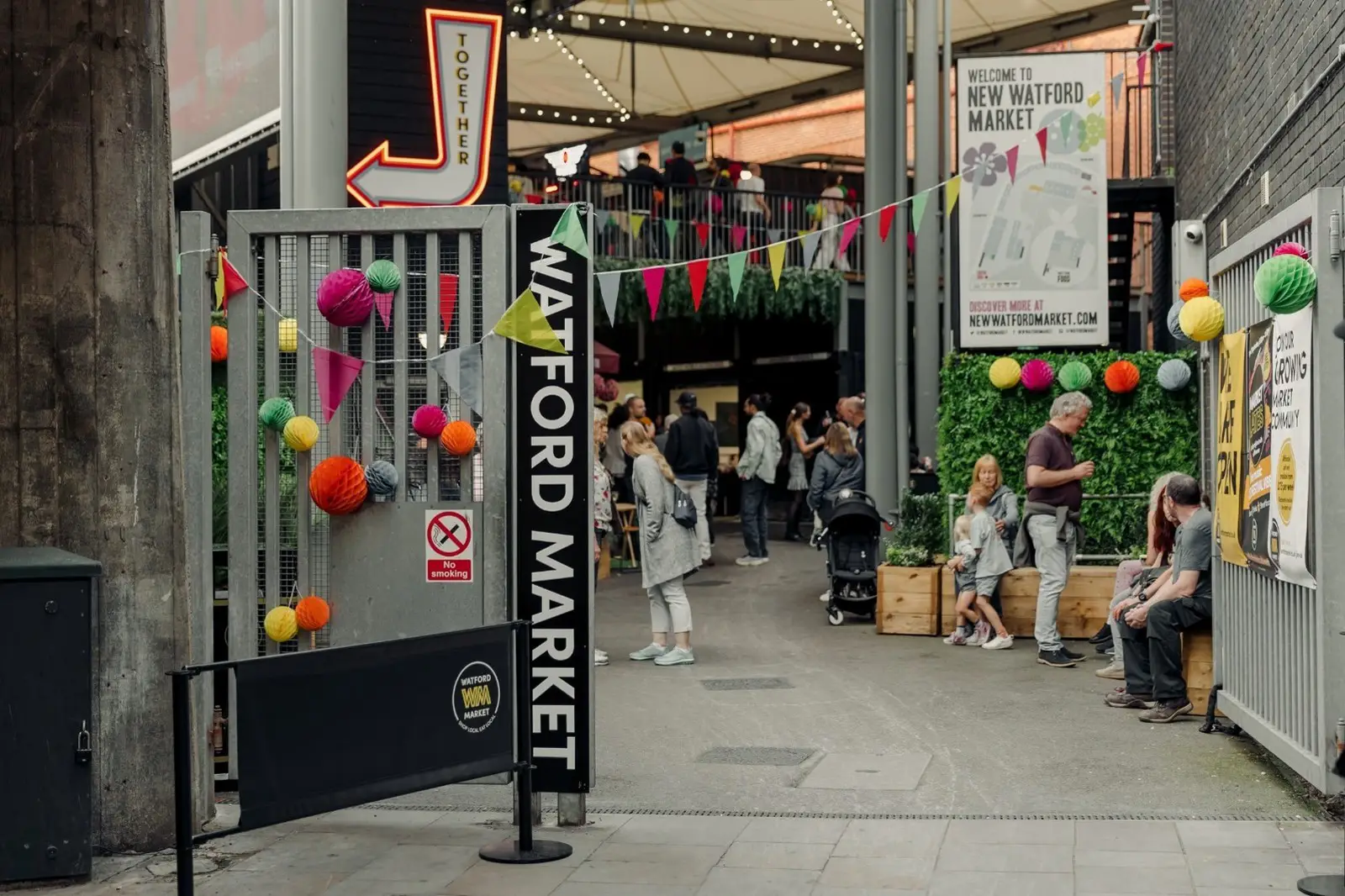 Entrance to Watford Market with colorful decorations and people walking and sitting nearby.