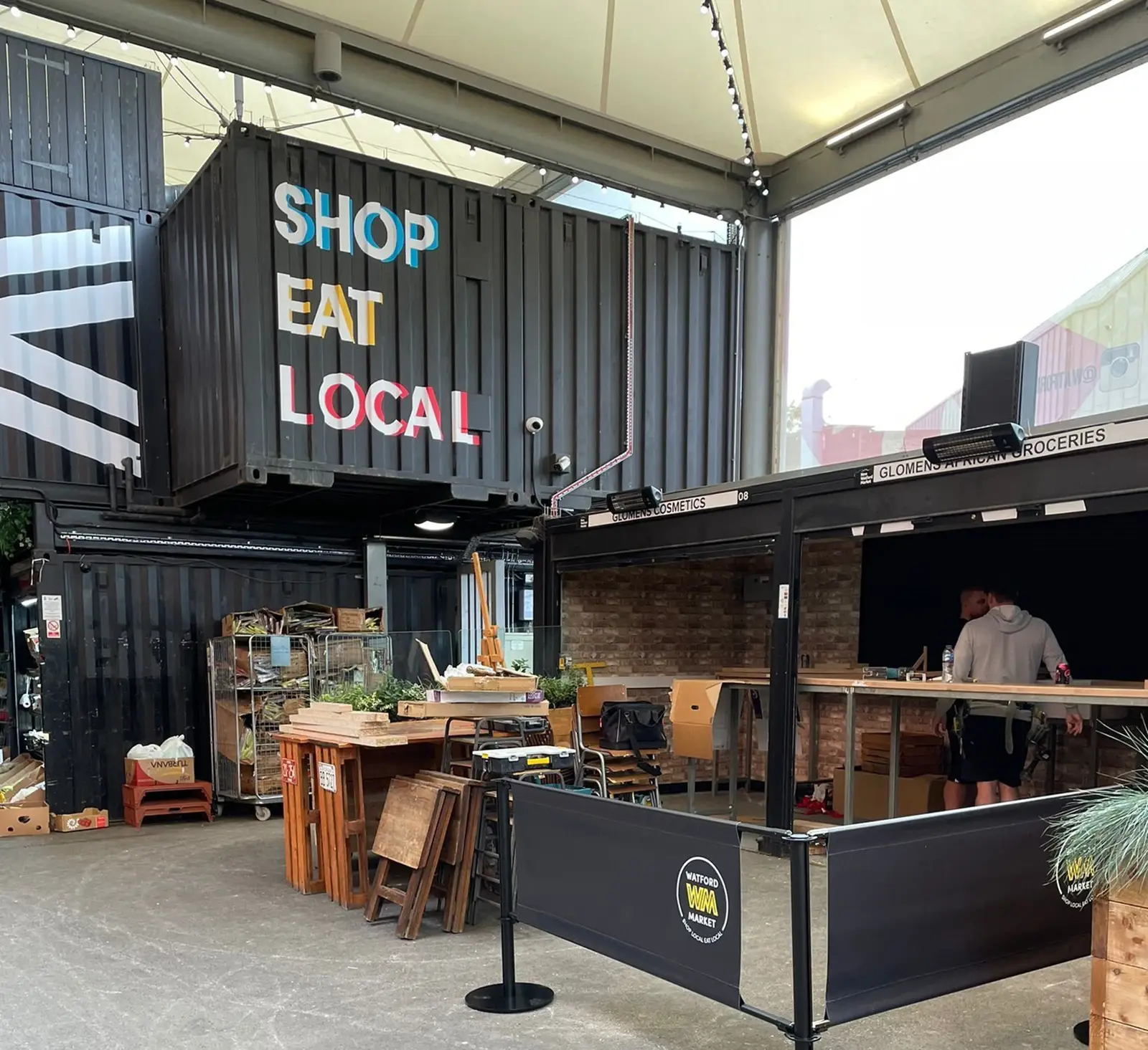 Outdoor market area with shipping containers, tables, and a sign reading "Shop Eat Local.