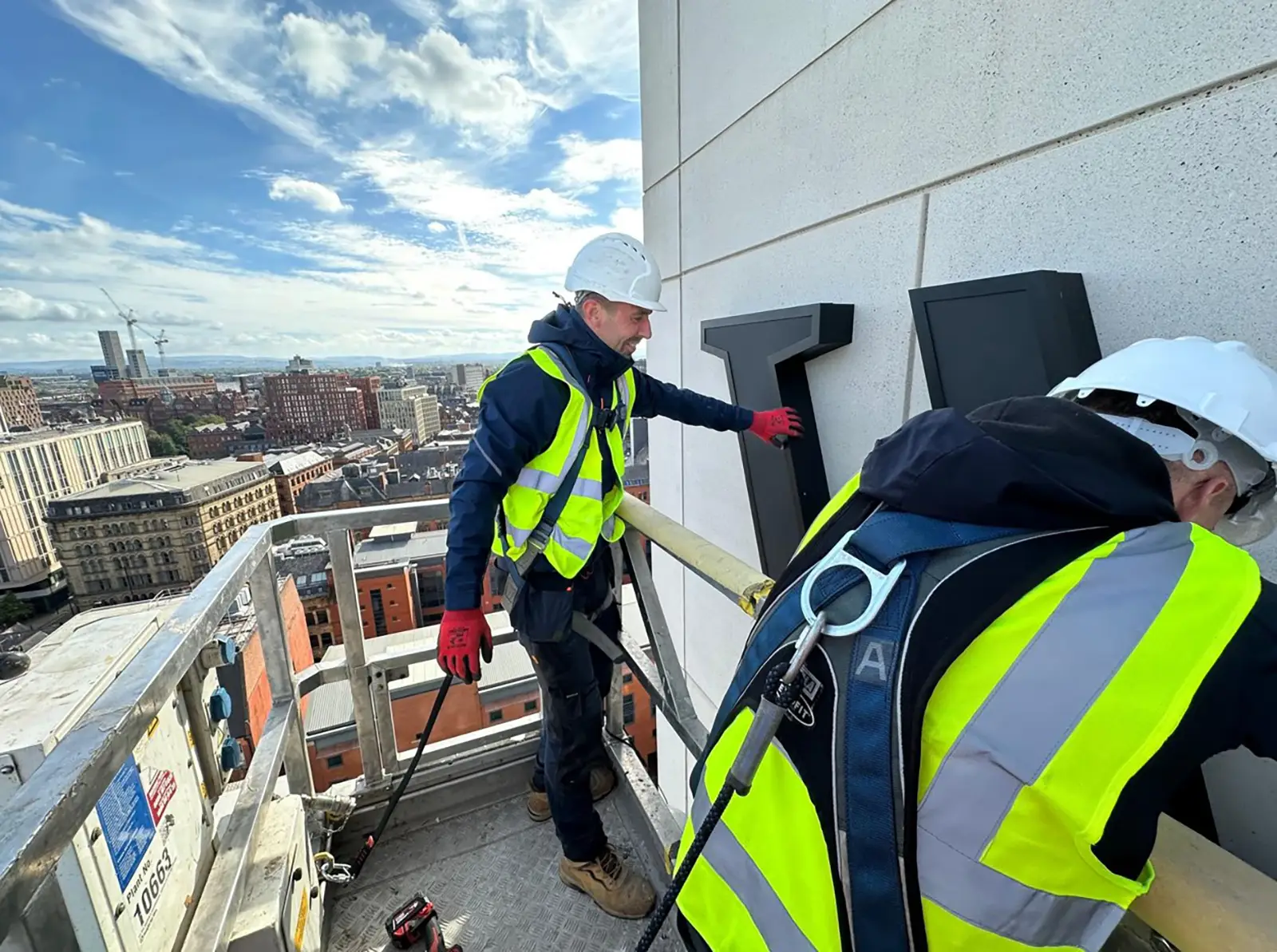 Two workers in safety gear install large letters on a building's exterior from a lift.