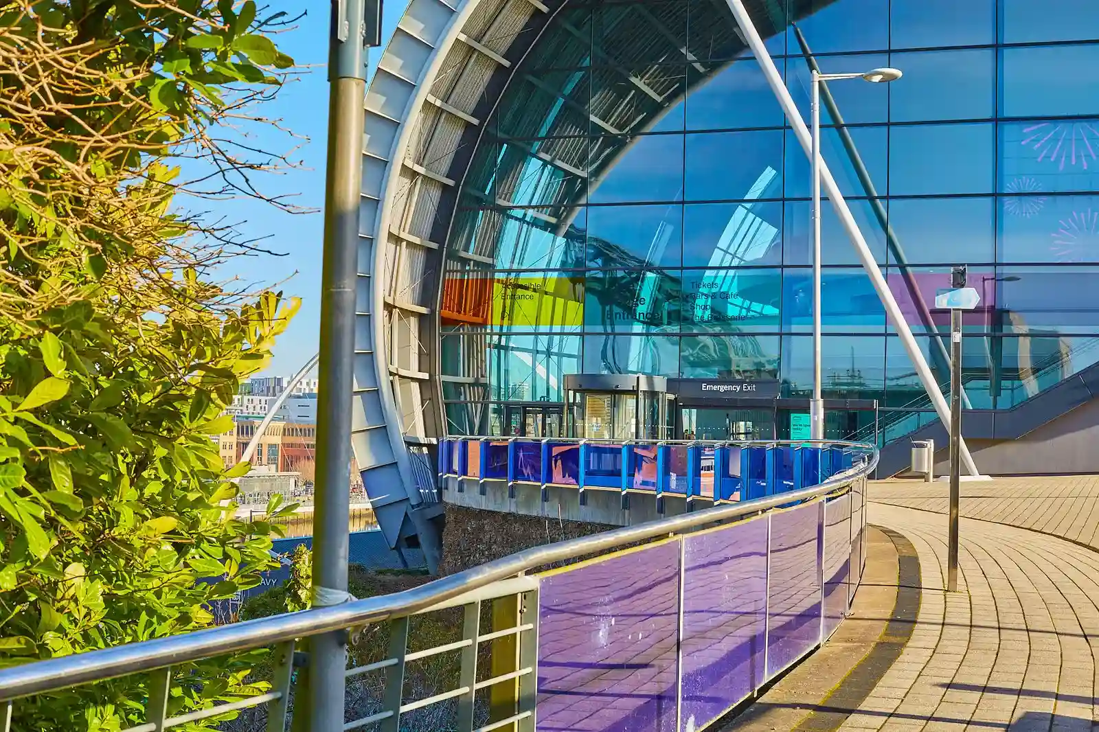 Modern glass building with colorful panels and a curved pathway, surrounded by greenery.