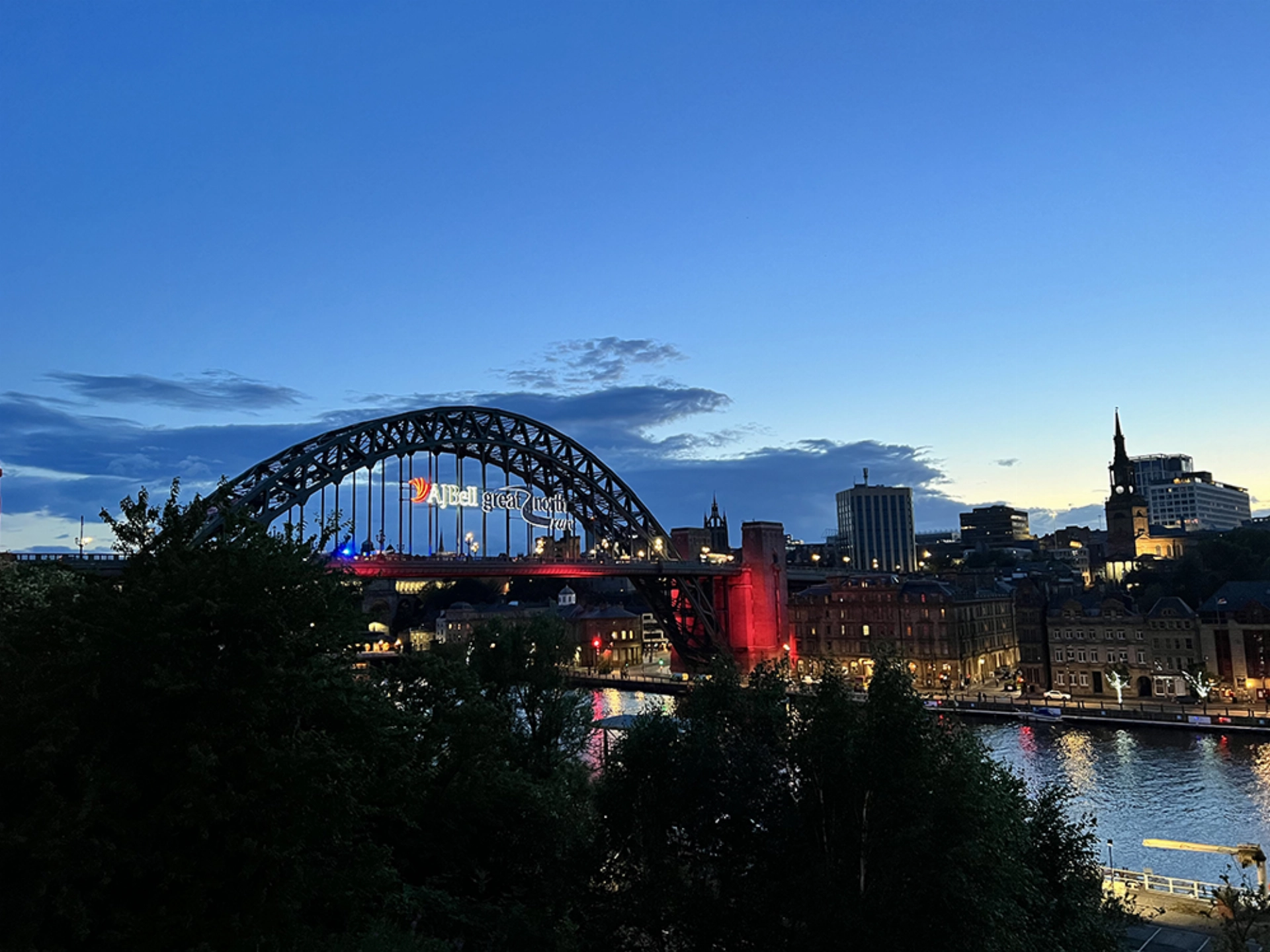 City skyline at dusk with a lit bridge over a river and illuminated buildings.