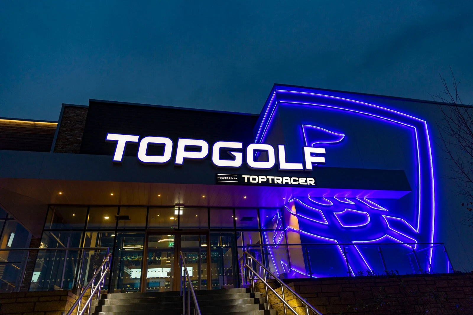 Topgolf building entrance with illuminated sign and neon blue logo at dusk.