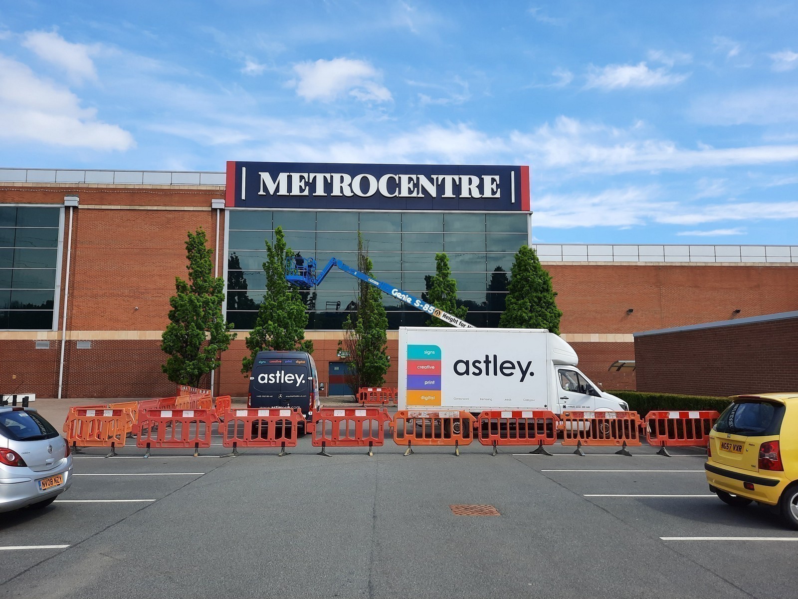 A white van and cherry picker in front of Metrocentre with orange barriers and trees.