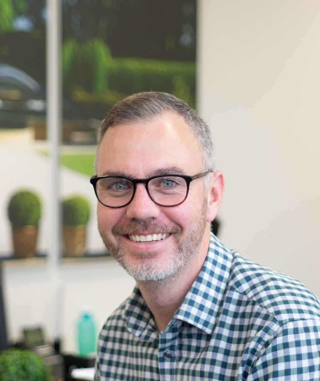 Man with glasses and a beard smiling, wearing a checkered shirt in an office setting.