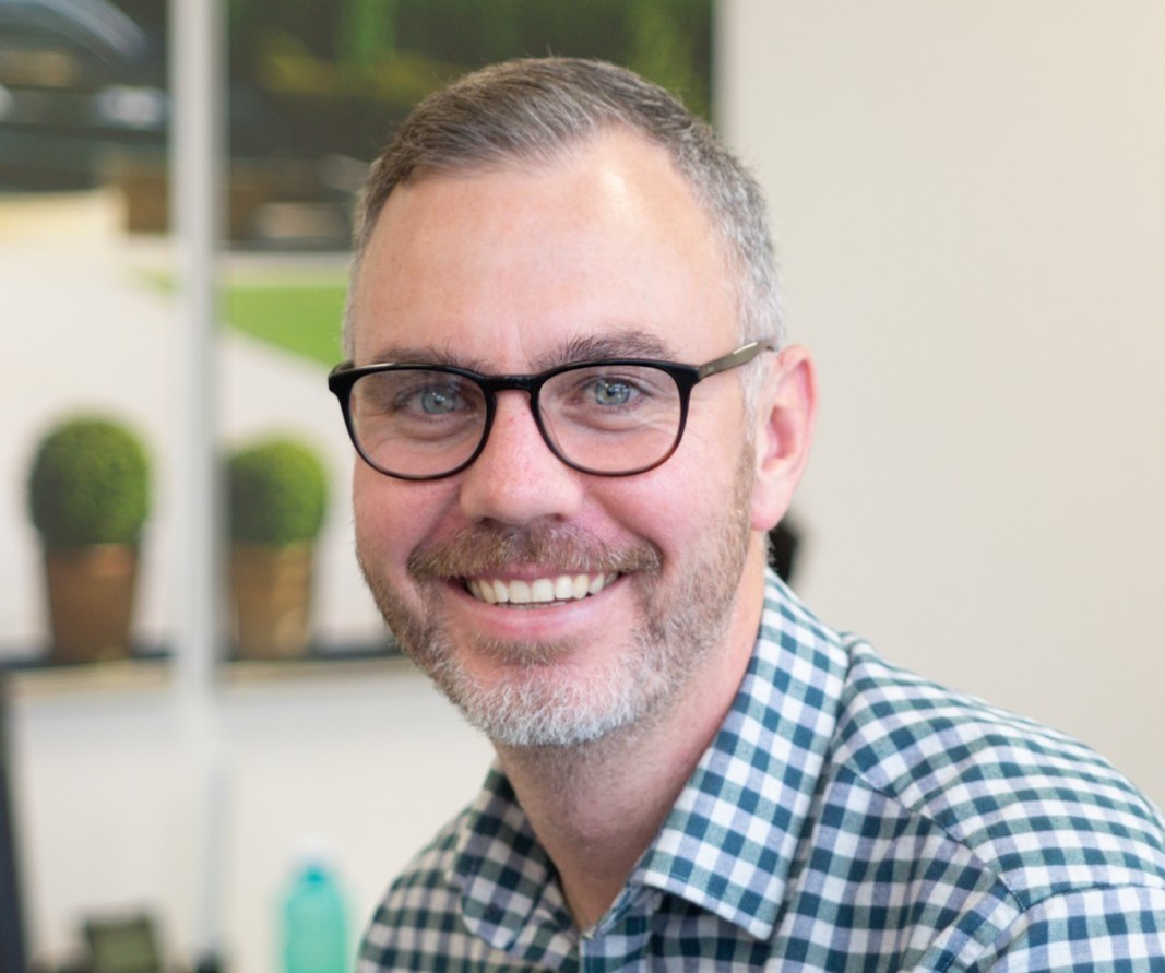 A man with glasses and a beard smiles, wearing a checkered shirt indoors.