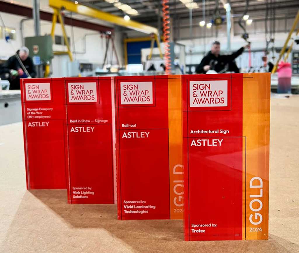 Four red and orange "Sign & Wrap Awards" plaques on a table in a workshop.