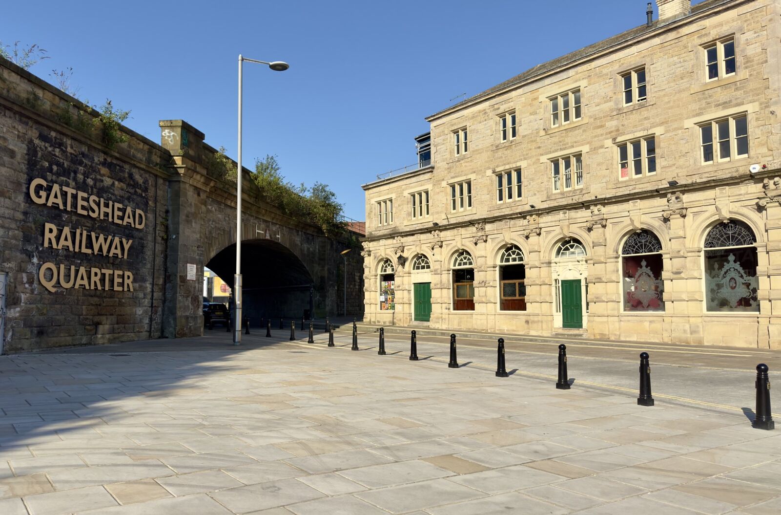 Stone buildings and an archway with "Gateshead Railway Quarter" sign under a clear blue sky.