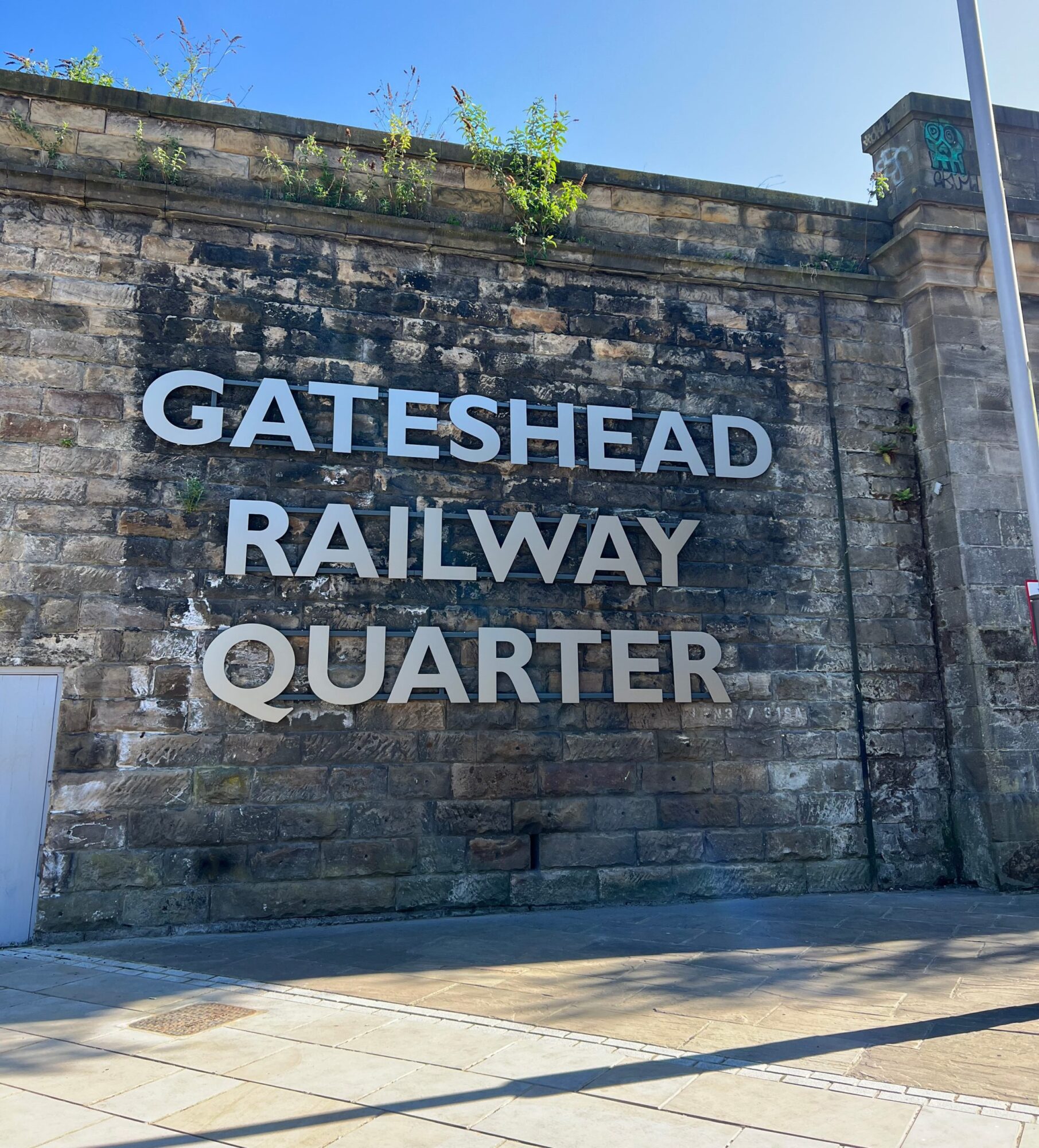 Stone wall with large letters spelling "GATESHEAD RAILWAY QUARTER" and some greenery on top.