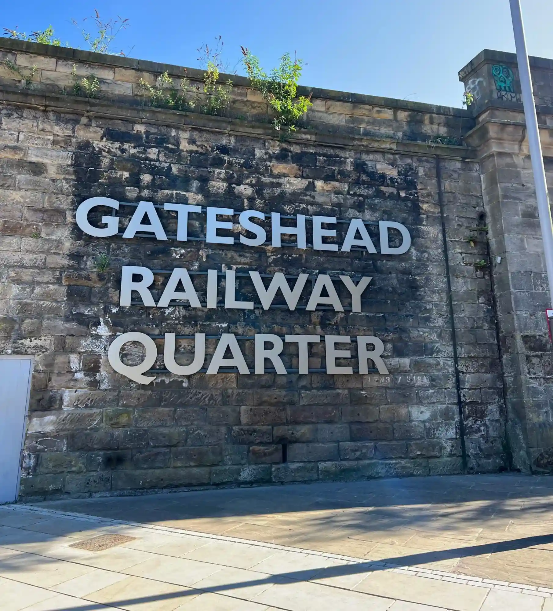Stone wall with large letters spelling "GATESHEAD RAILWAY QUARTER" and some greenery on top.