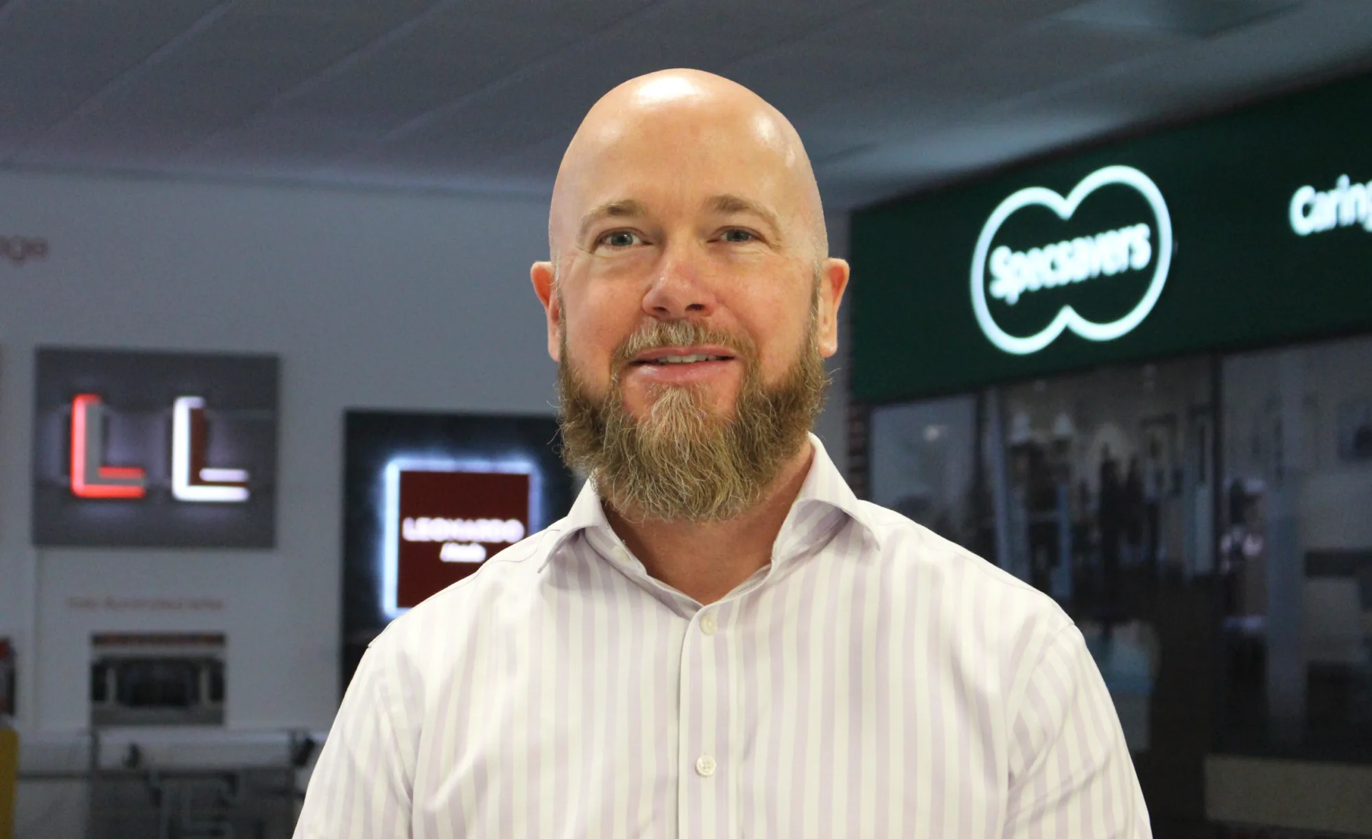 A bald man with a beard smiles in front of a Specsavers store sign.