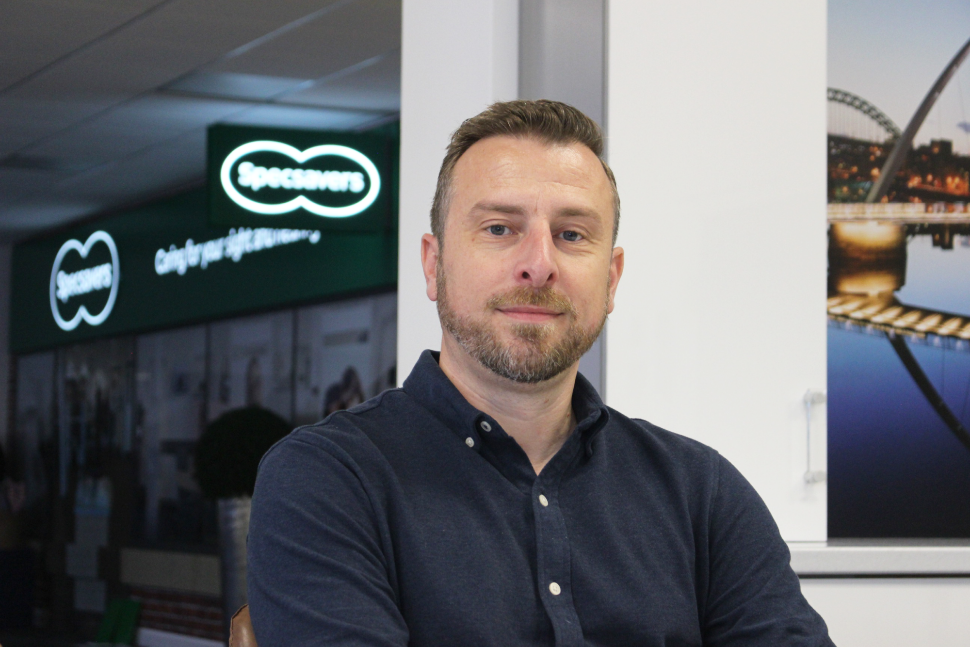 A man with a beard and blue shirt stands in front of a Specsavers store.