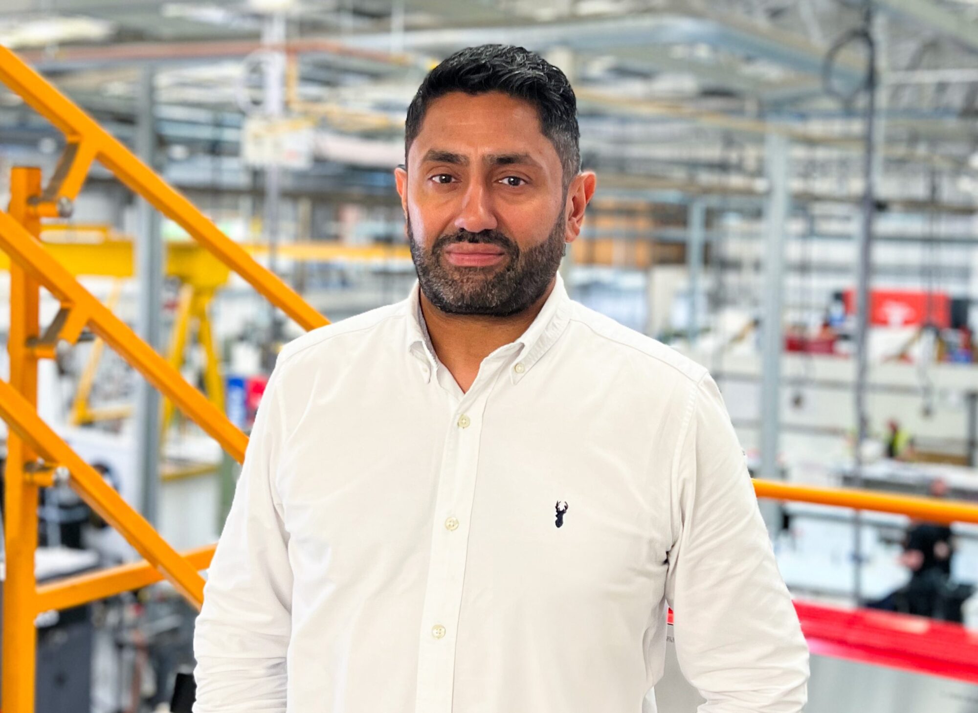 A man in a white shirt stands in an industrial setting with machinery in the background.