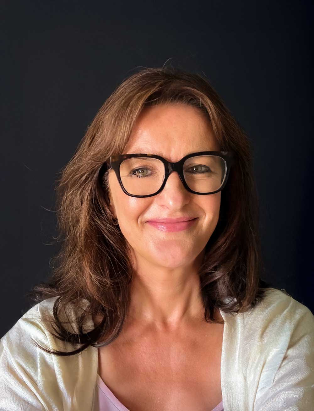 A person with glasses and long hair smiles against a dark background.