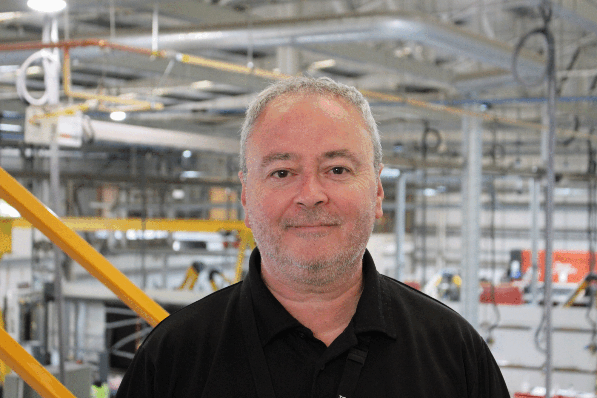 A man in a black polo shirt stands in an industrial setting, with a background of machinery, pipes, and yellow safety railings in a factory or warehouse environment.