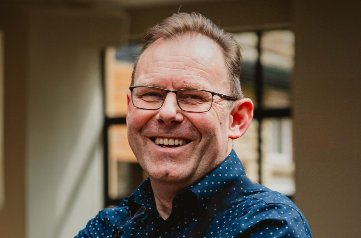 A man with glasses and a blue polka dot shirt smiles indoors near a window.