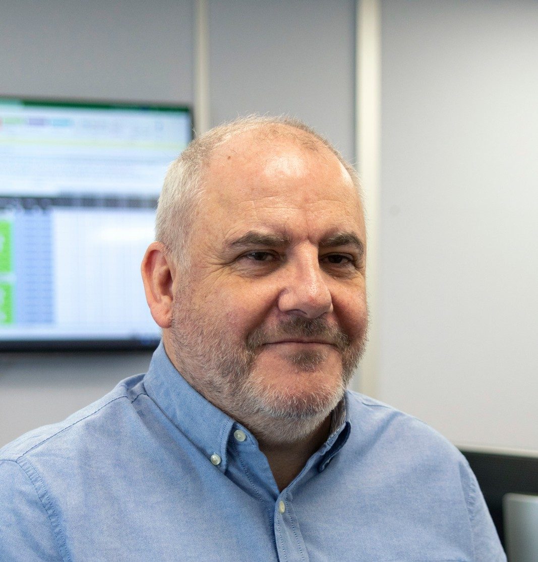 A man with a beard and blue shirt sits in an office with a screen behind.
