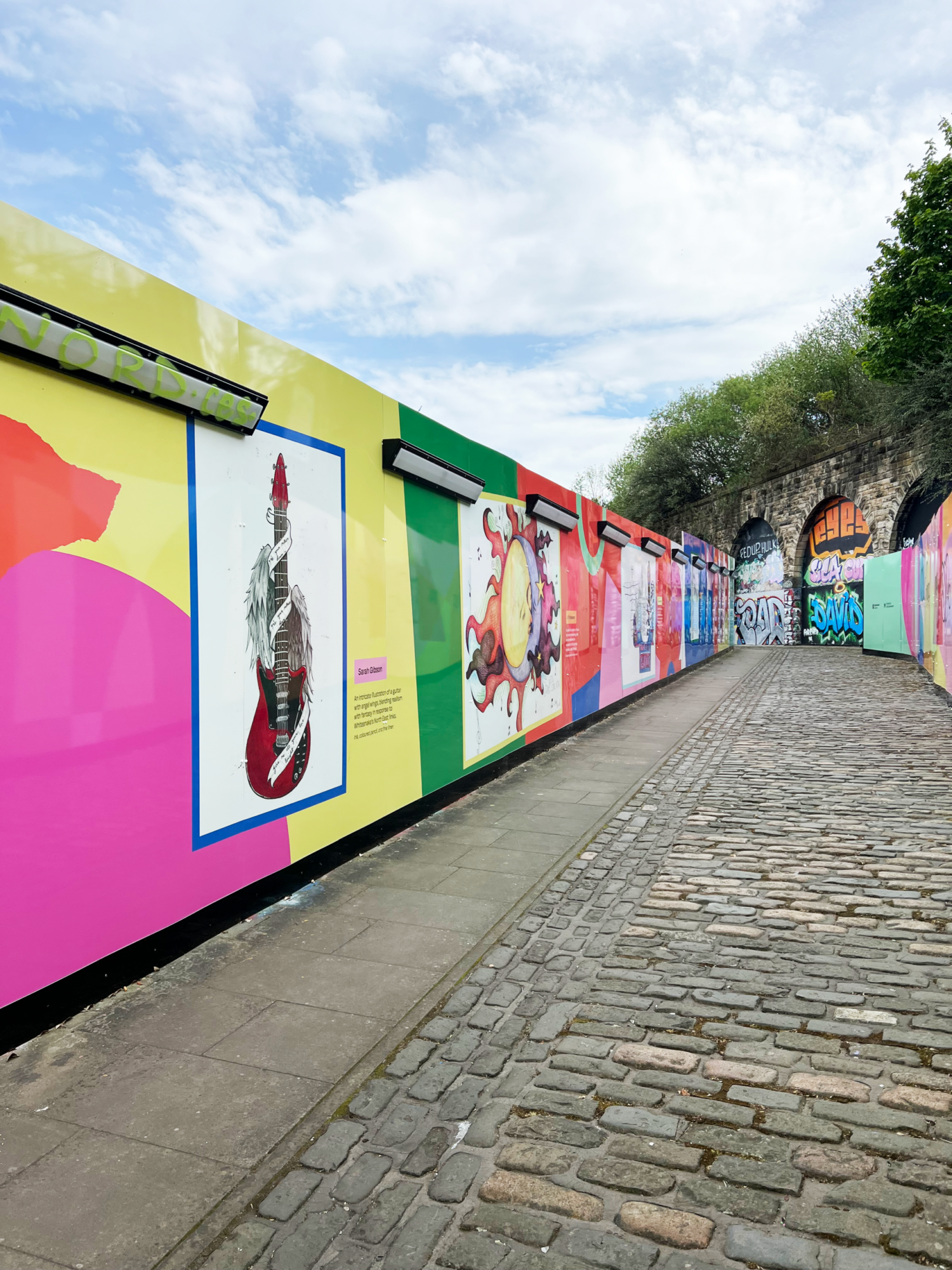Colorful murals line a cobblestone path with a stone archway and trees in the background.