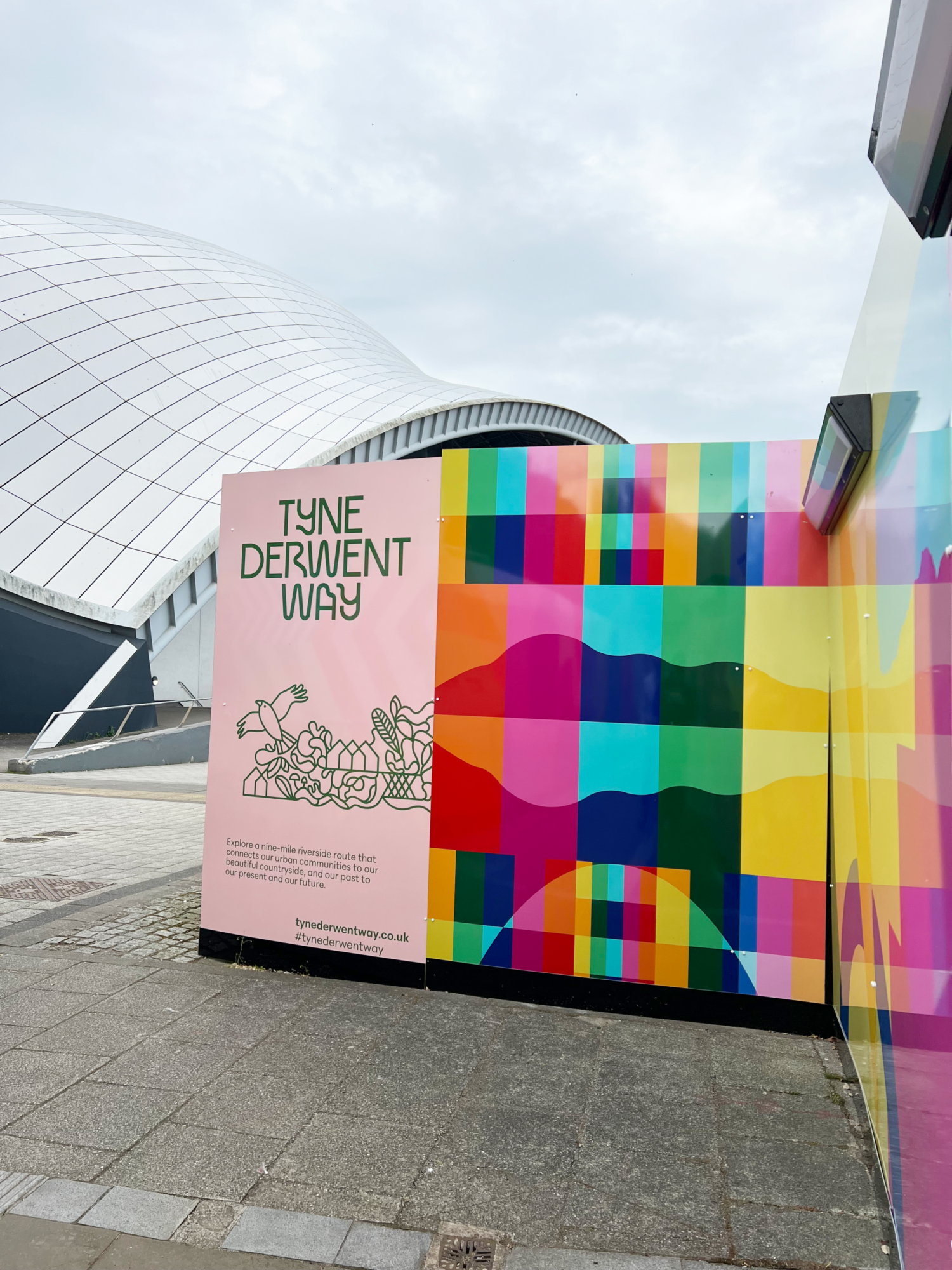 Colorful mural and "Tyne Derwent Way" sign near a modern, curved building under a cloudy sky.