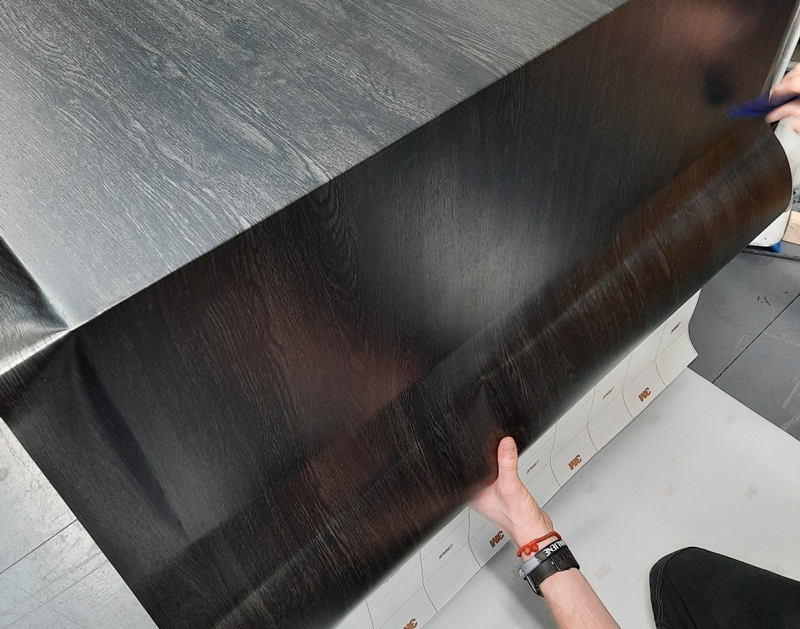 Hands applying a dark wood-patterned adhesive sheet onto a surface with a gray wood texture.