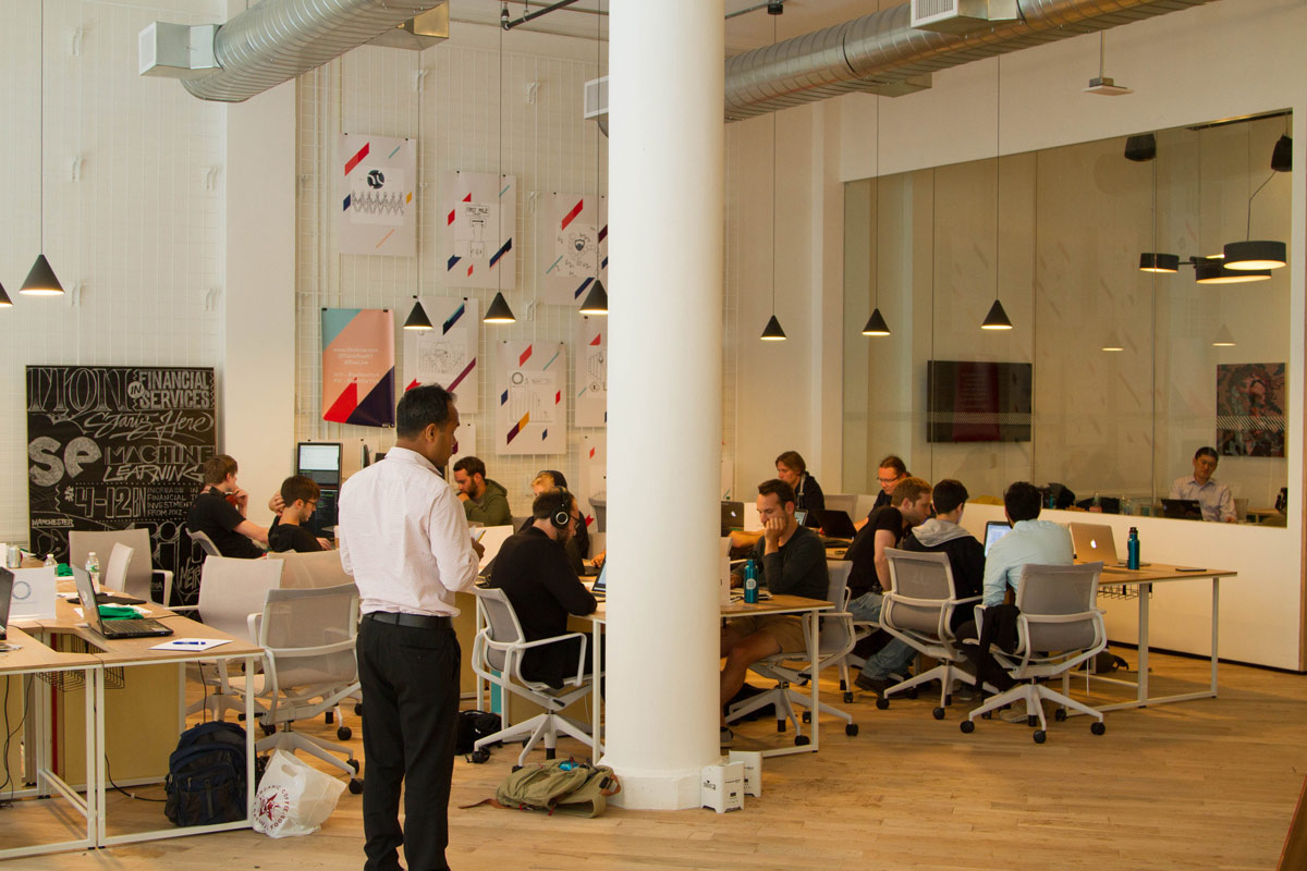 Open office space with modern industrial design; features groups of people collaborating at desks with laptops and monitors. White pillar in foreground, wall art displays geometric patterns. Overhead lights and exposed ductwork create a contemporary ambiance.