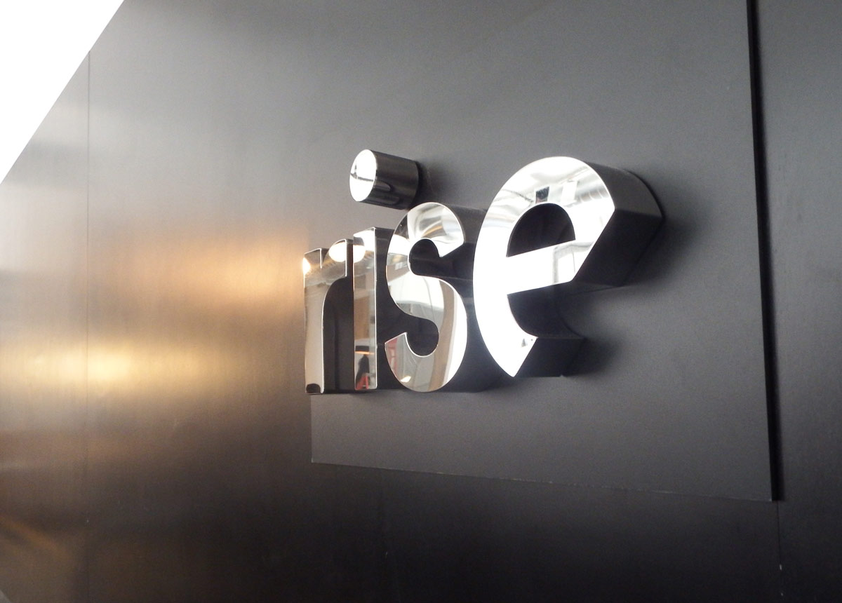 The word "rise" in shiny, metallic letters is mounted on a black wall.